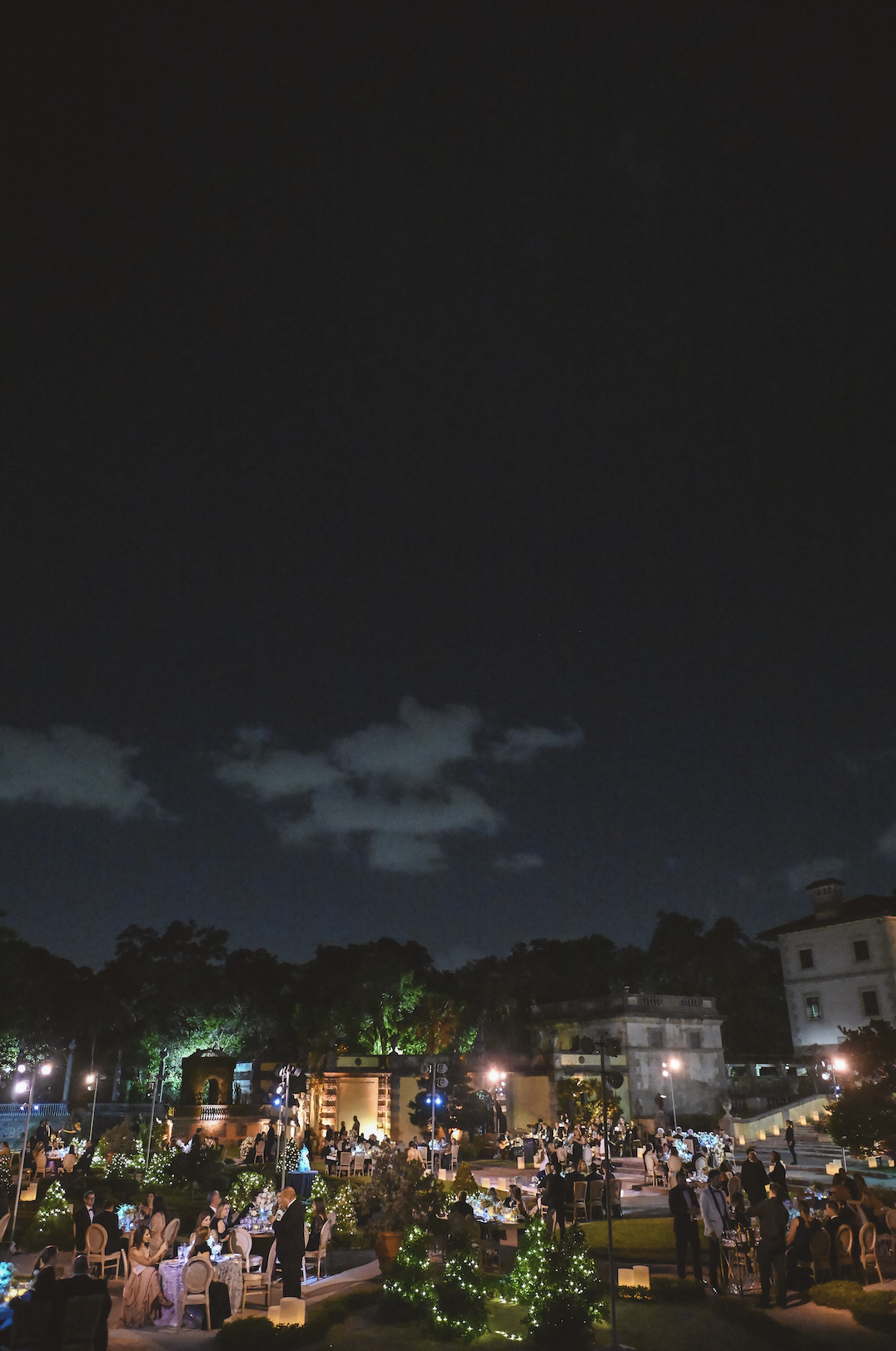 Nighttime outdoor celebration with tables of people, decorative lights, and historic buildings in the background under a dark sky.