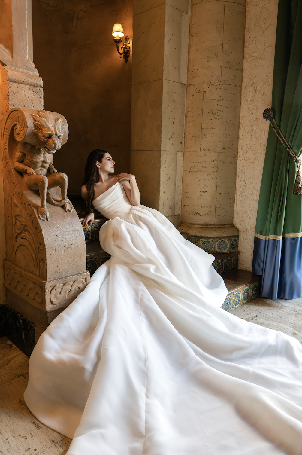 A woman in a white wedding gown sits on a stone bench near a large green curtain and decorative tile steps, with a lion statue head beside her, in a luxurious interior setting with warm lighting.
