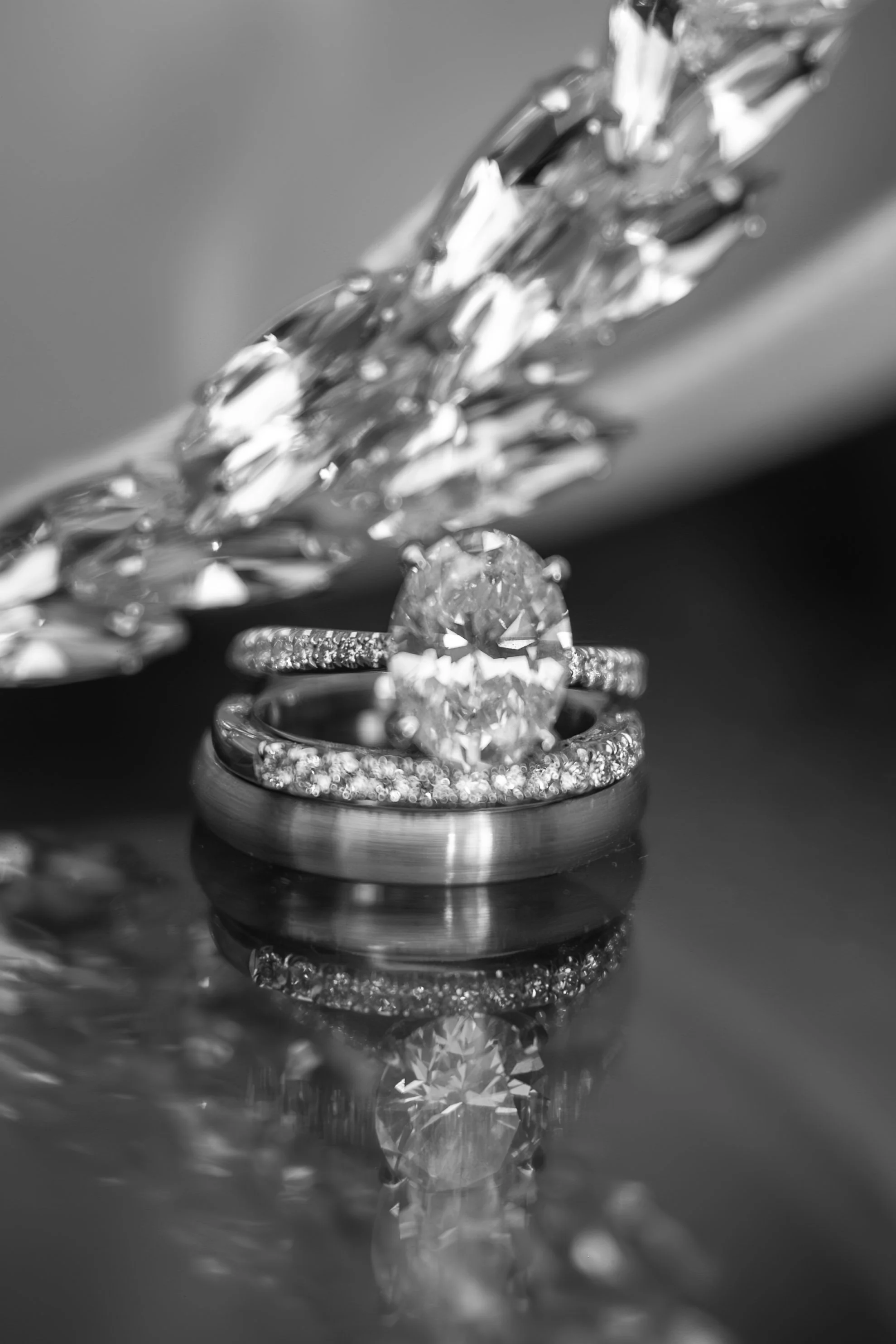 Close-up of a diamond engagement ring and wedding band with intricate designs, placed on a reflective surface with a blurred background.