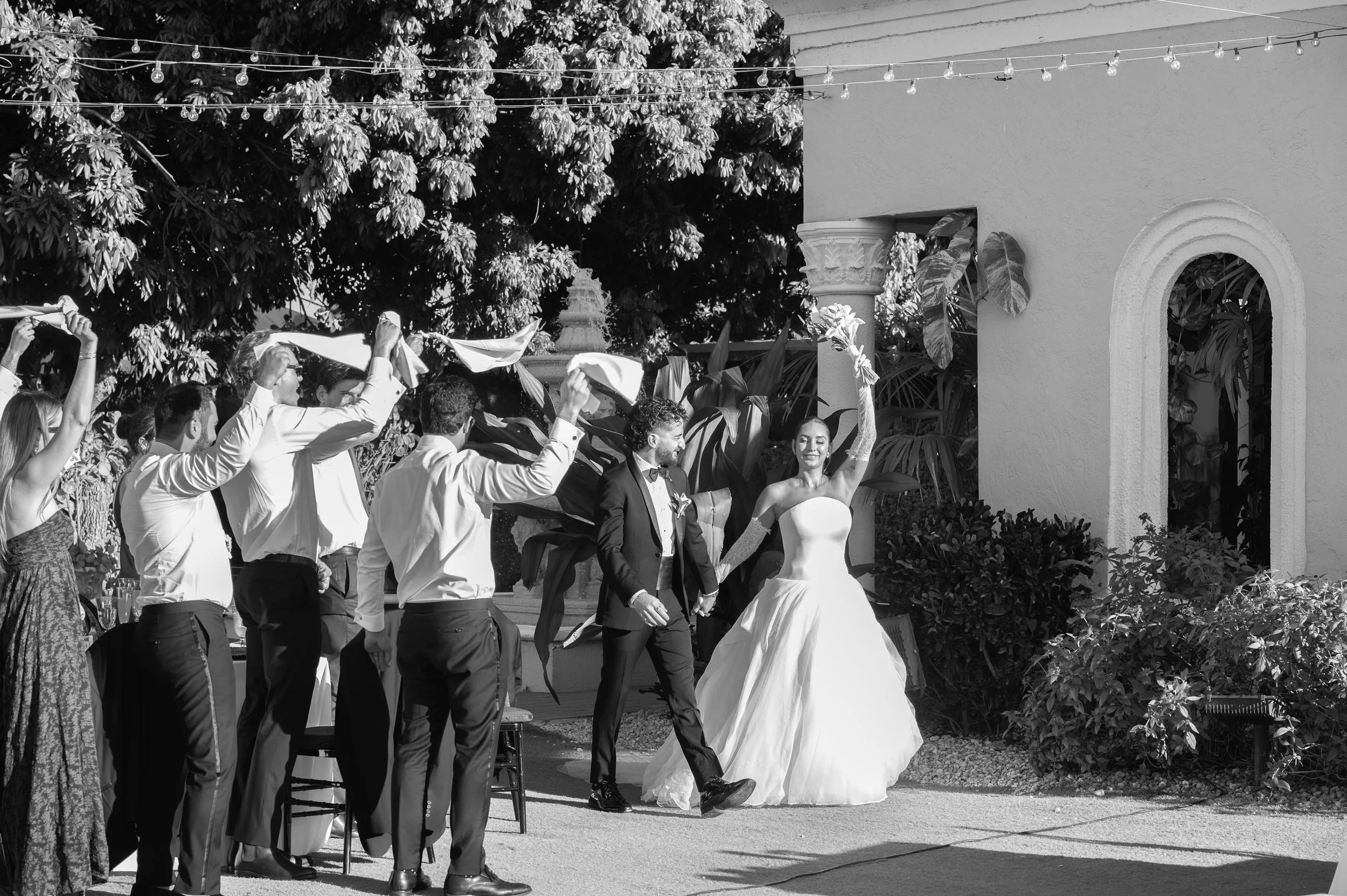 A wedding reception outdoors with people dancing, a bride in a white gown smiling, and a groom in a tuxedo, under string lights with lush greenery and a building wall in the background.