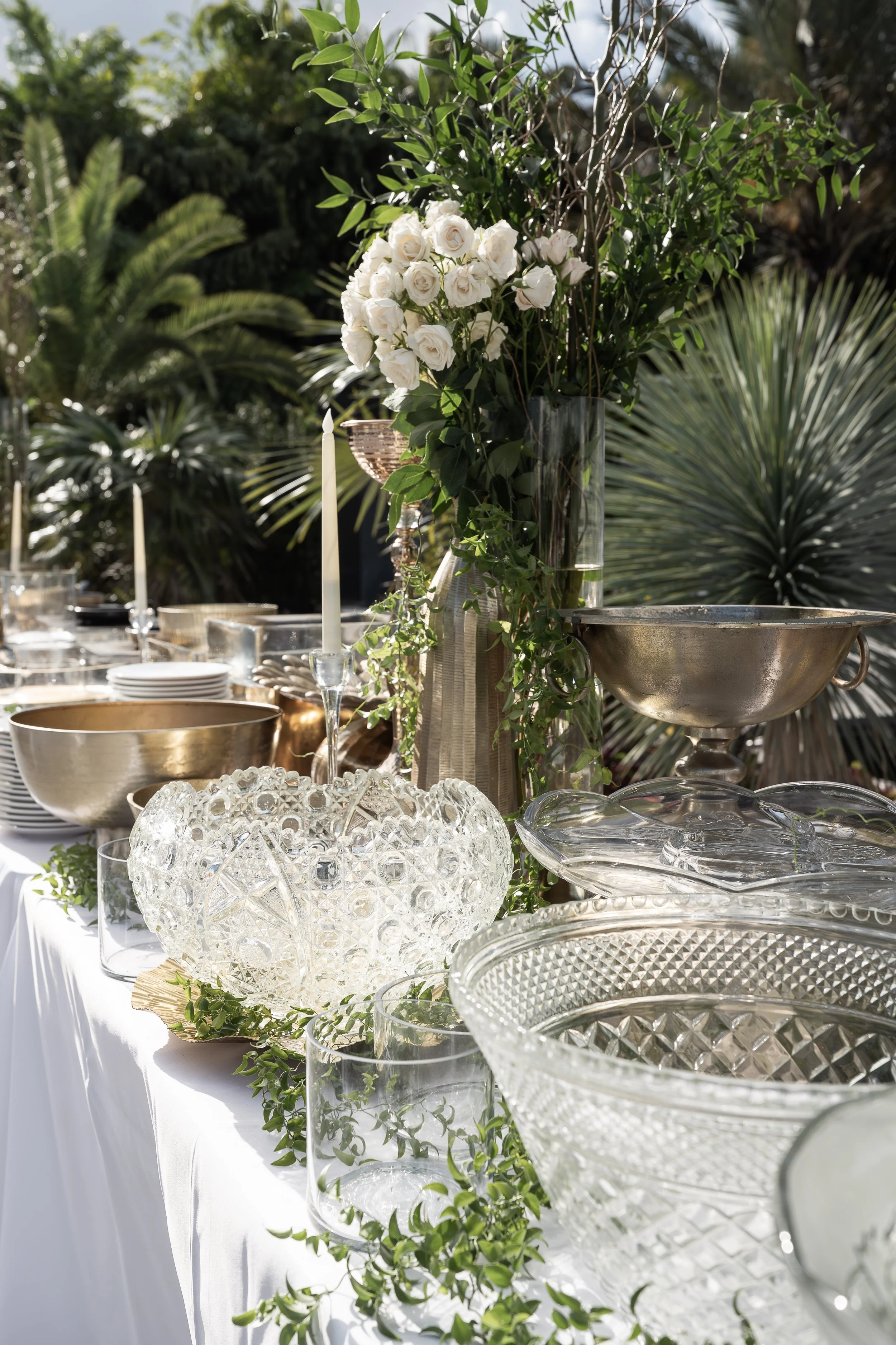 A table decorated with glass bowls, a large vase of white roses and greenery, and candles, set outdoors with tropical plants in the background.