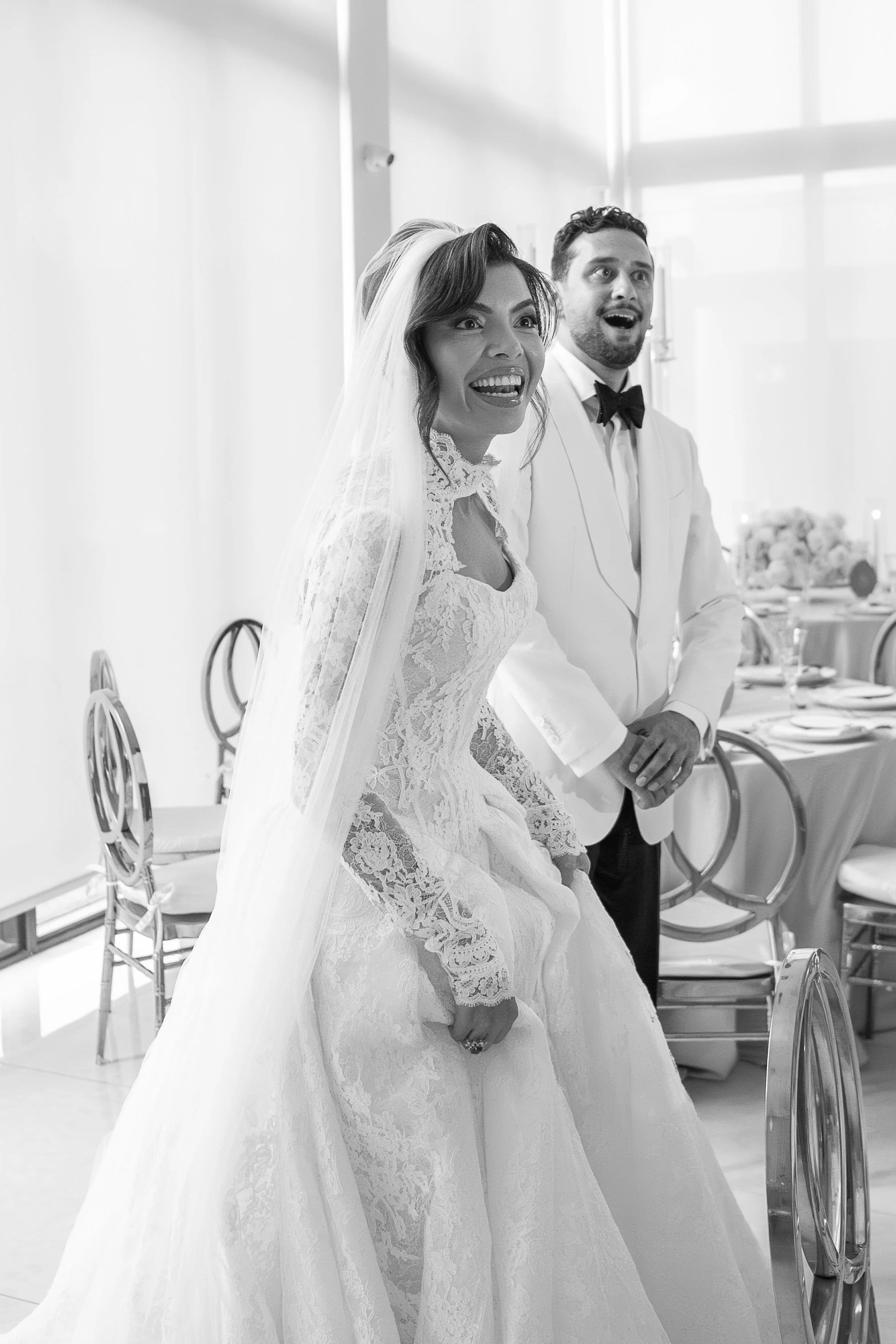 Black and white photo of a bride and groom at their wedding reception, both with surprised expressions, standing next to each other in front of a decorated dining table.