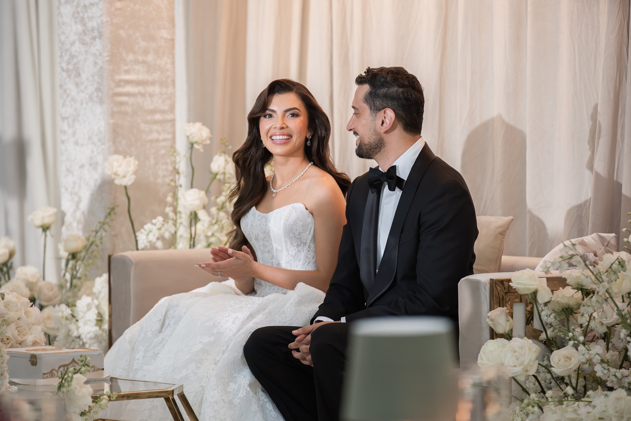 A bride and groom sitting together during their wedding ceremony, surrounded by white flowers and elegant decor.