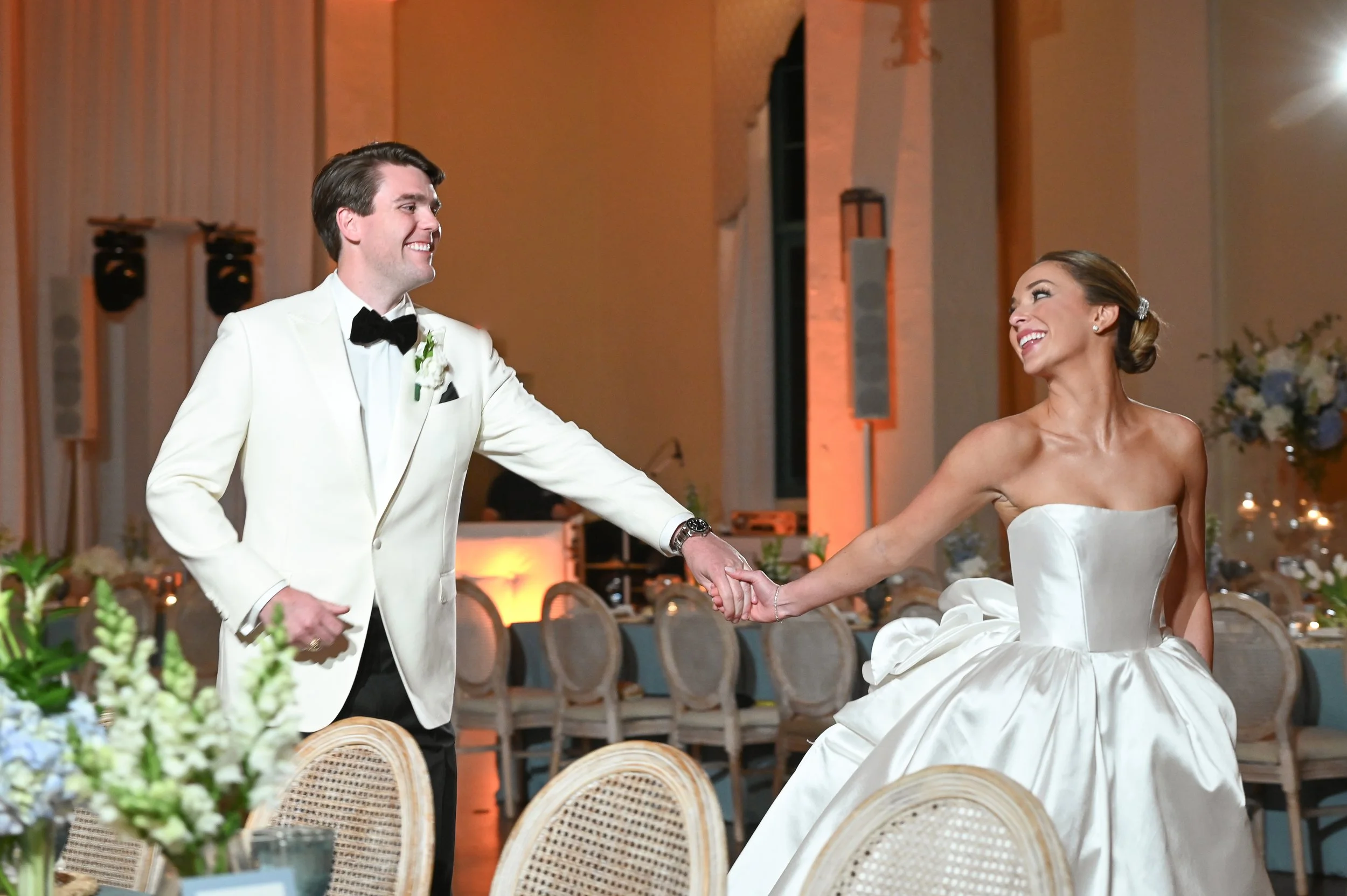 A newlywed couple dancing at their wedding reception, smiling and holding hands in a beautifully decorated indoor venue with floral arrangements and tables.