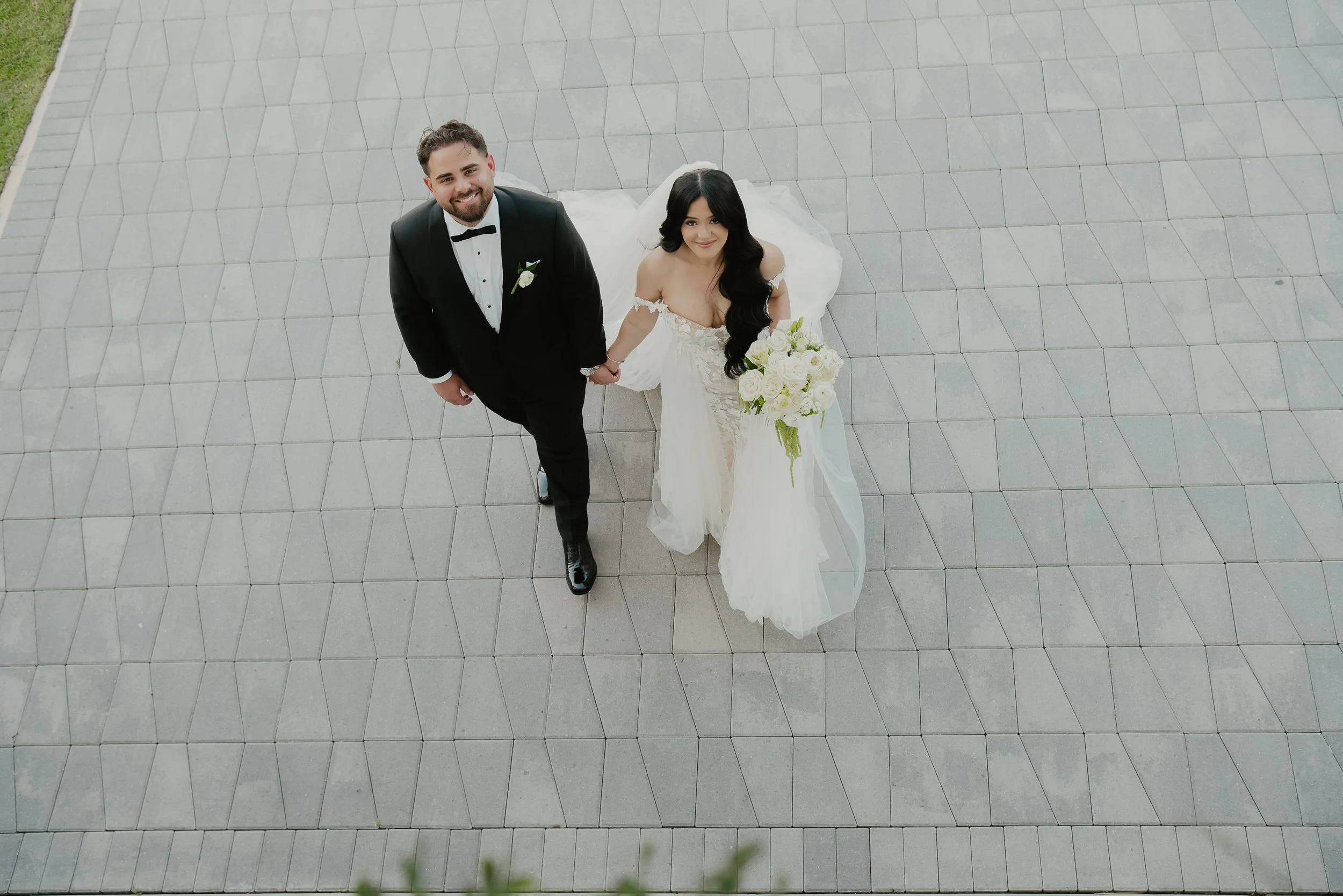 A bride and groom looking up at the camera, holding hands, standing on a tiled outdoor area. The groom is in a black tuxedo and bowtie, the bride is in a white wedding dress holding a bouquet of white roses, with her long black hair in loose waves.
