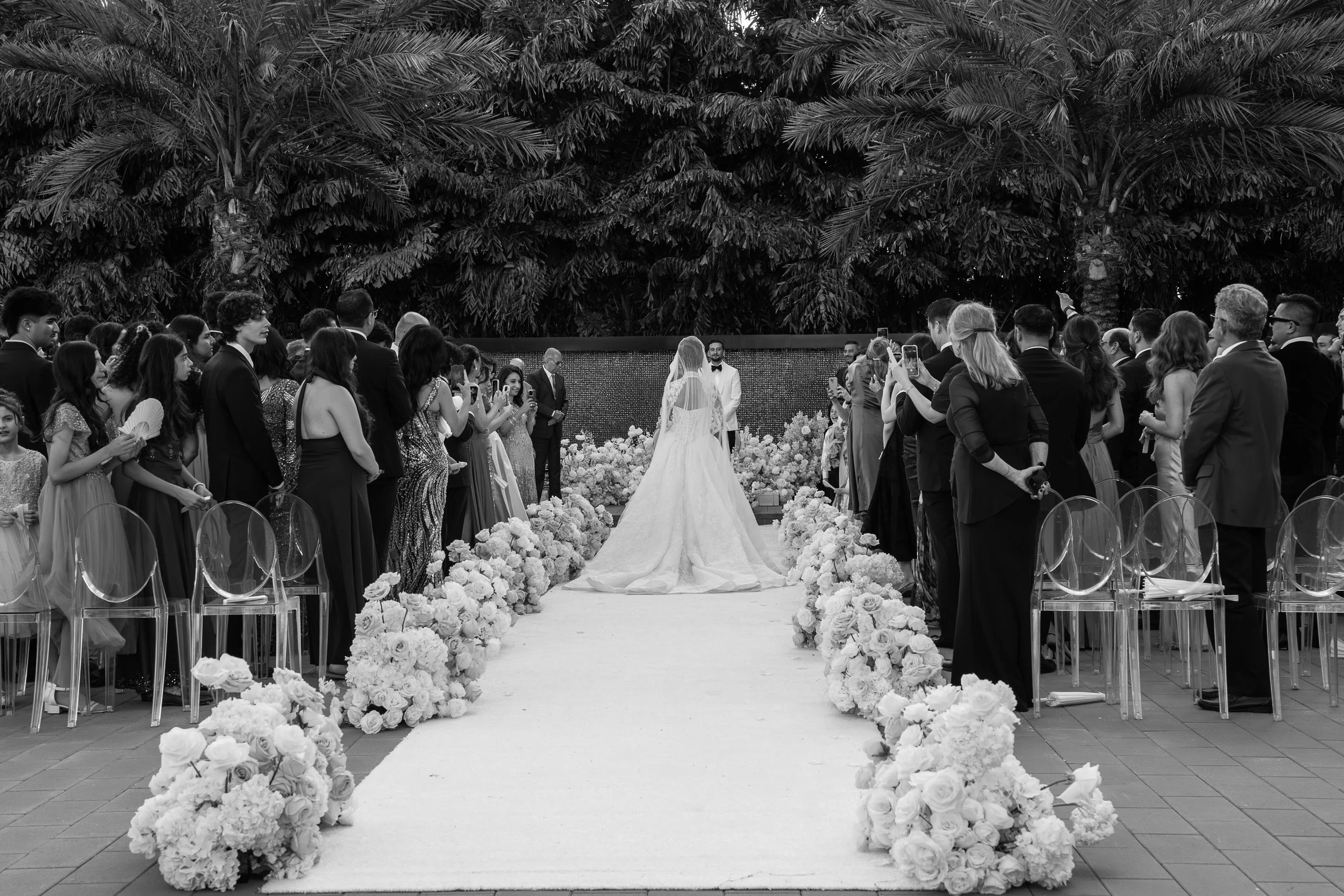 A wedding ceremony outdoors with a bride and groom standing at the altar, surrounded by guests, with large palm trees in the background.