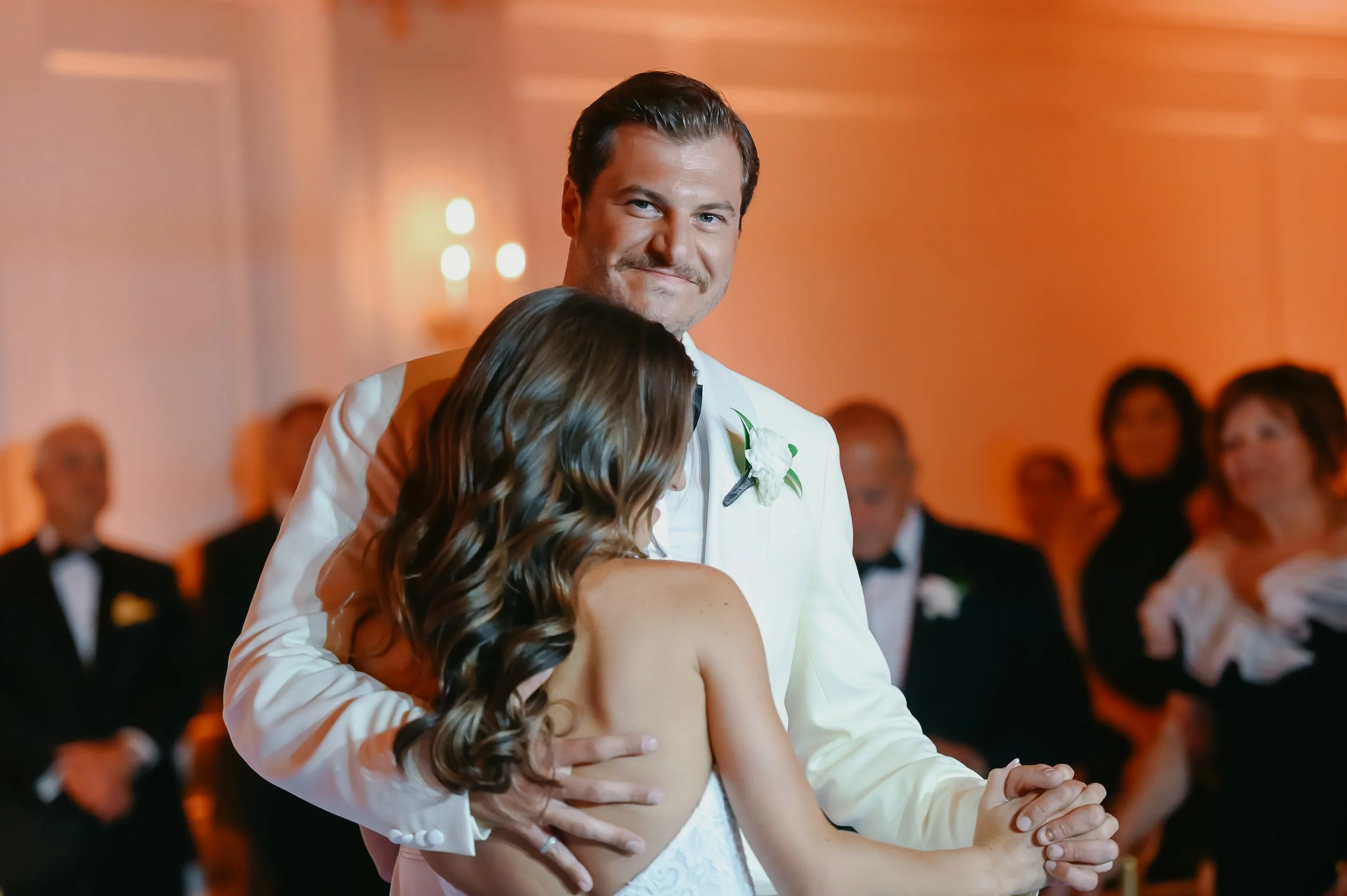 A man and woman share a dance at a formal event, with the man wearing a white tuxedo and the woman in a white gown. They are surrounded by seated guests in formal attire, and the background is warm with orange lighting.