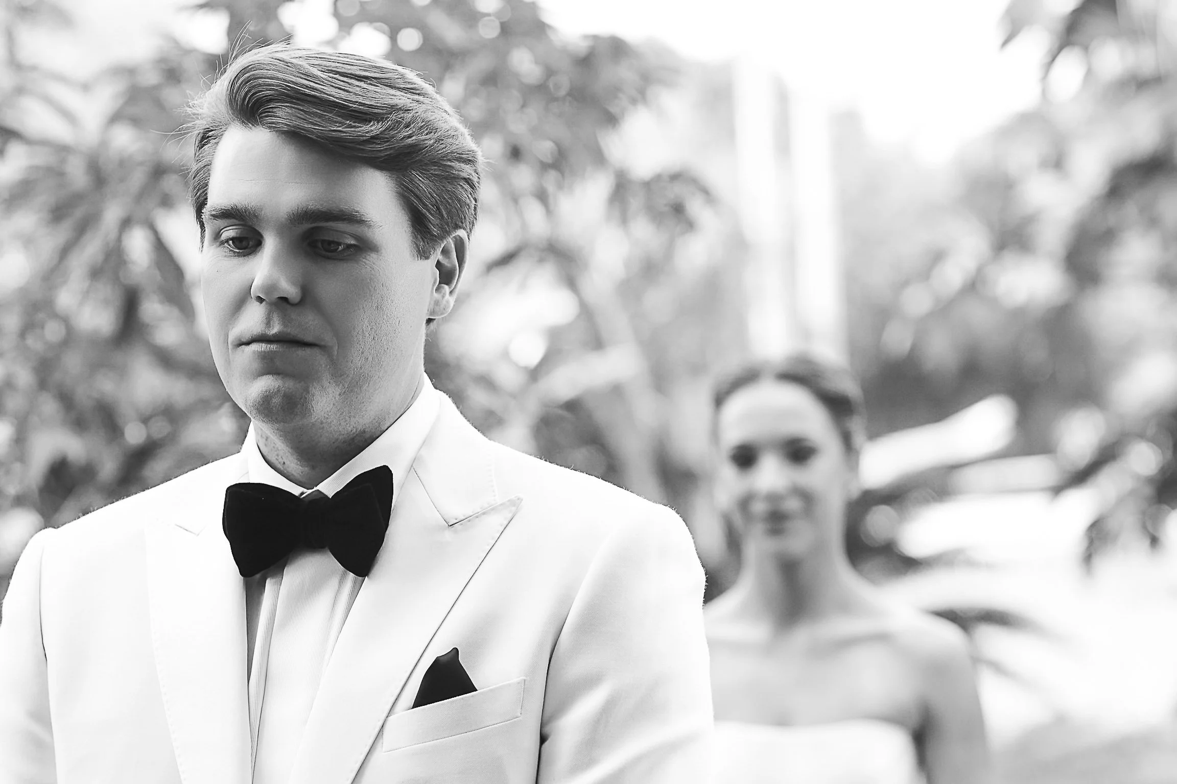 A young man dressed in a white tuxedo with a black bow tie, looking down thoughtfully. He has styled hair and is outdoors with blurred trees in the background.