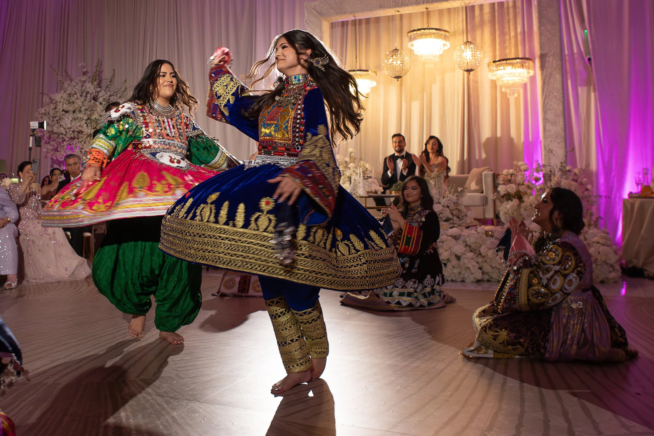 Two women performing a traditional Indian dance at a wedding reception, wearing colorful, embroidered attire, with guests clapping and watching in a decorated hall with chandeliers and floral arrangements in the background.
