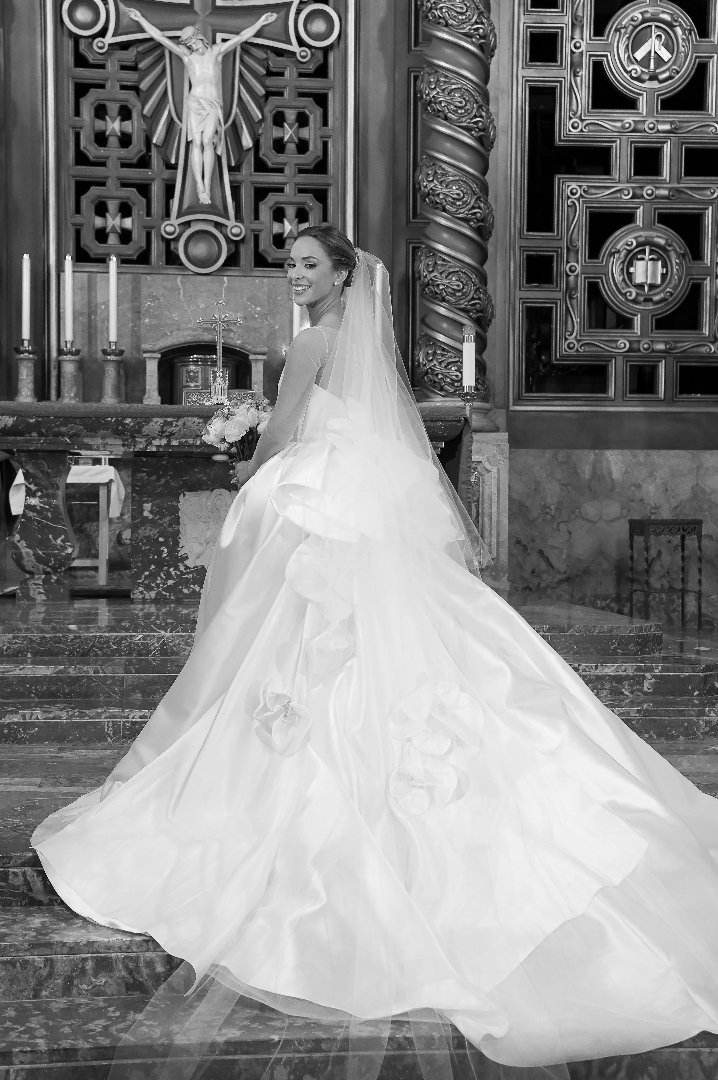 A bride in a wedding gown smiling and holding a bouquet, standing inside a church with religious decor and a crucifix behind her.
