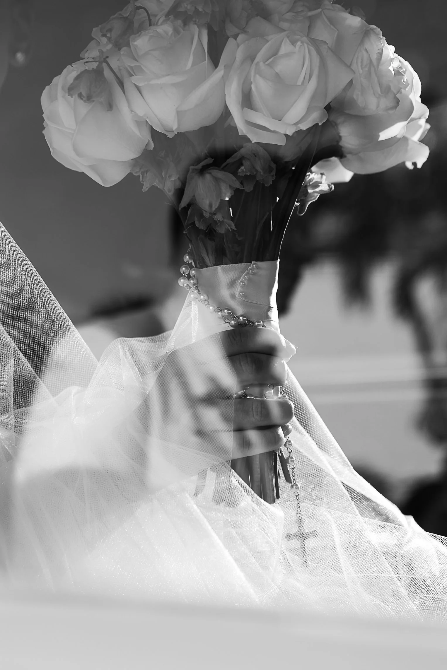 A bouquet of white roses wrapped in white tulle and a pearl necklace, held in someone's hand.