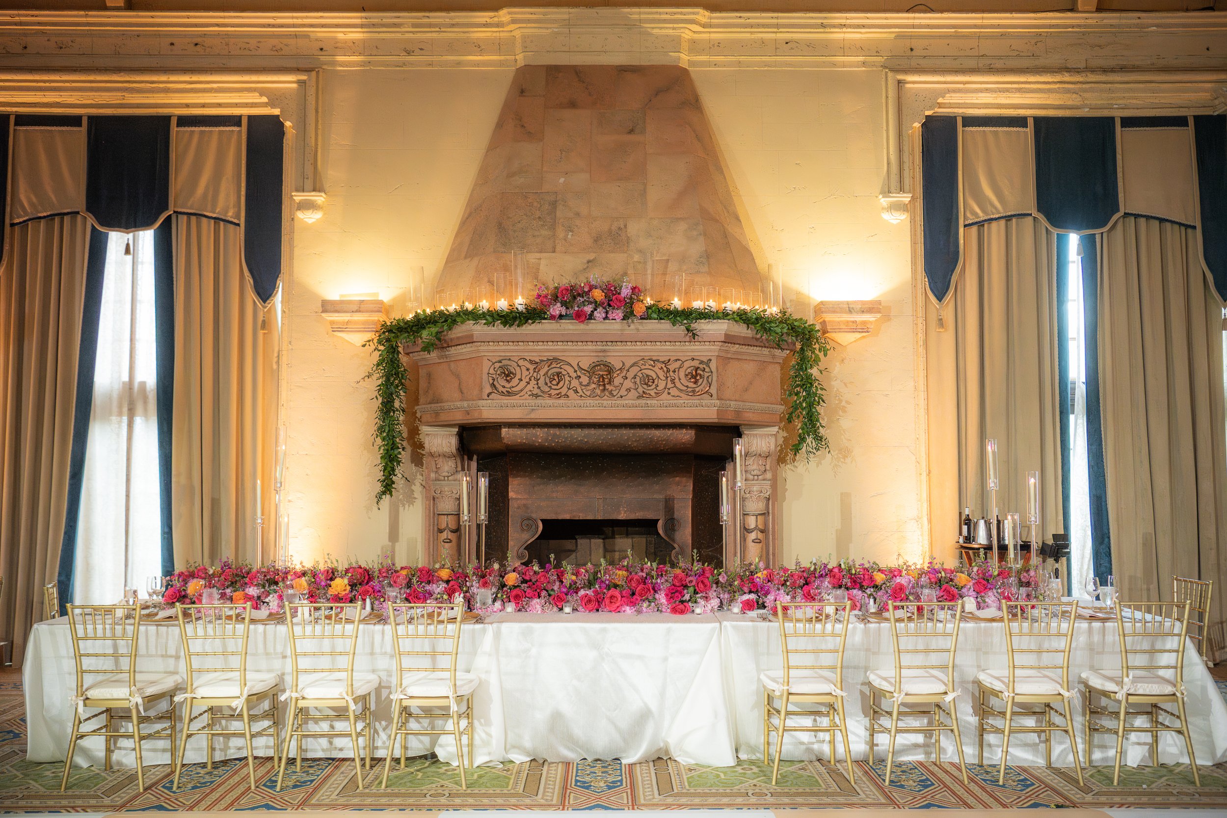 Elegant banquet table in front of a fireplace decorated with pink and orange flowers and greenery, set in a room with tall curtains and ornate molding, ambient lighting.