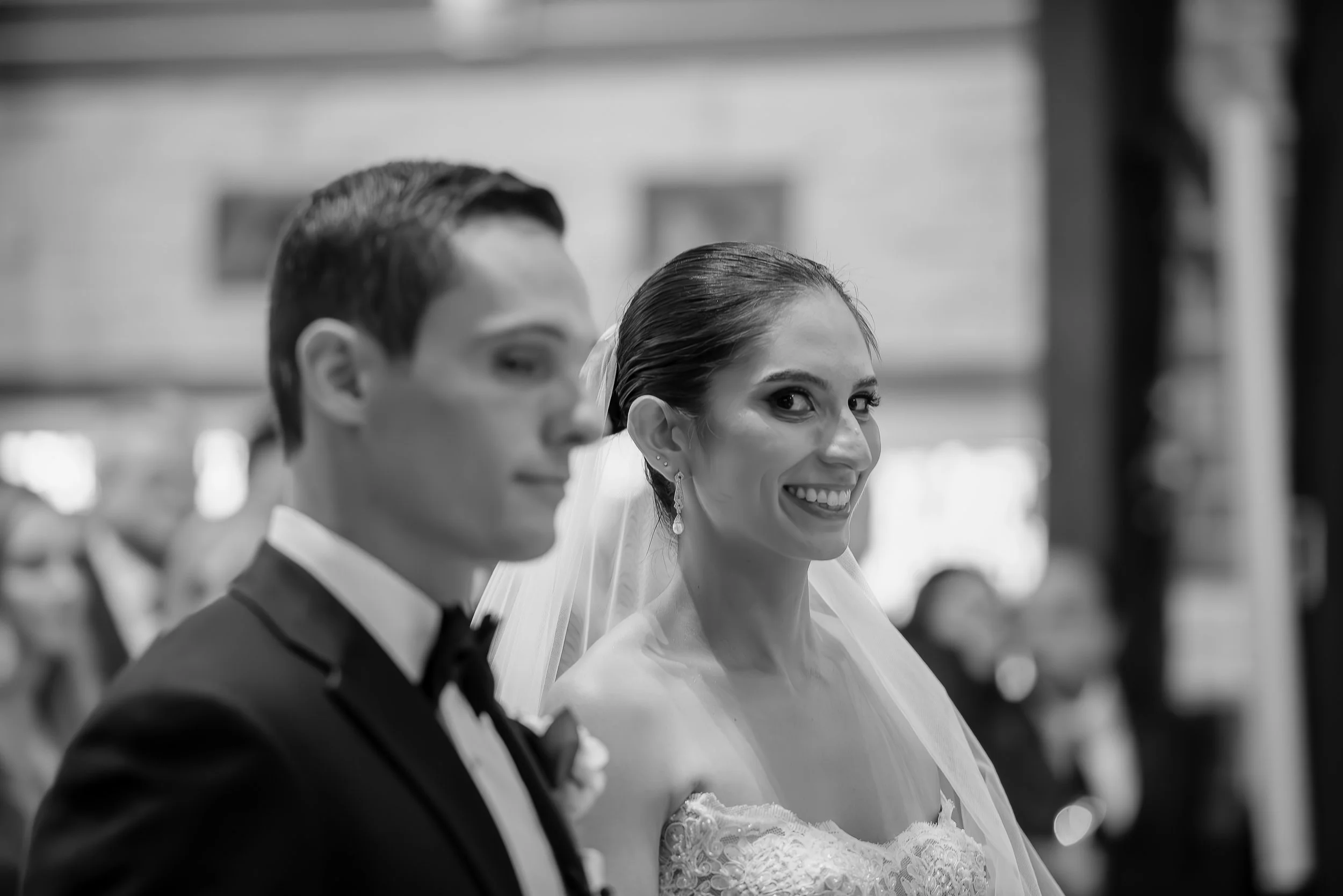 A bride and groom standing side by side during a wedding ceremony, with the bride smiling and the groom looking ahead, both dressed in formal wedding attire.