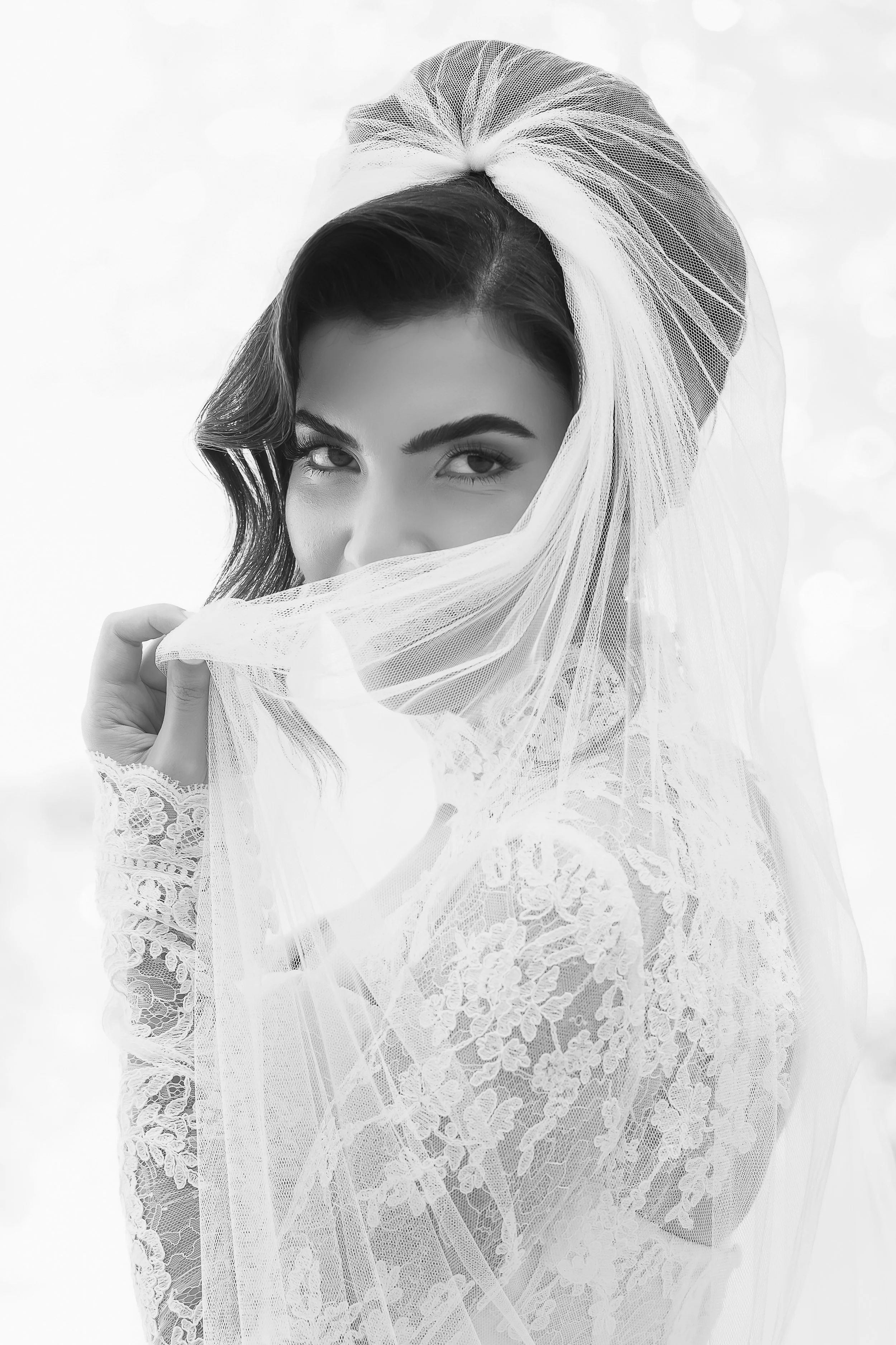 Black and white photo of a woman with dark hair and eye makeup, partially covered with a lace and tulle fabric, looking at the camera.