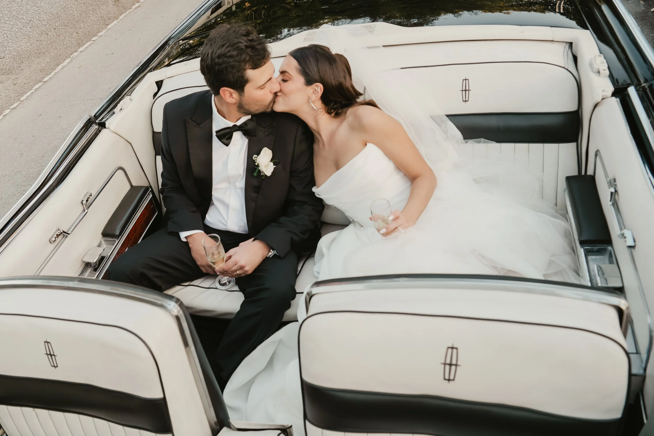A newly married couple kissing while sitting in the backseat of a white vintage car, with the bride wearing a wedding dress and veil and the groom in a black tuxedo, holding champagne glasses.