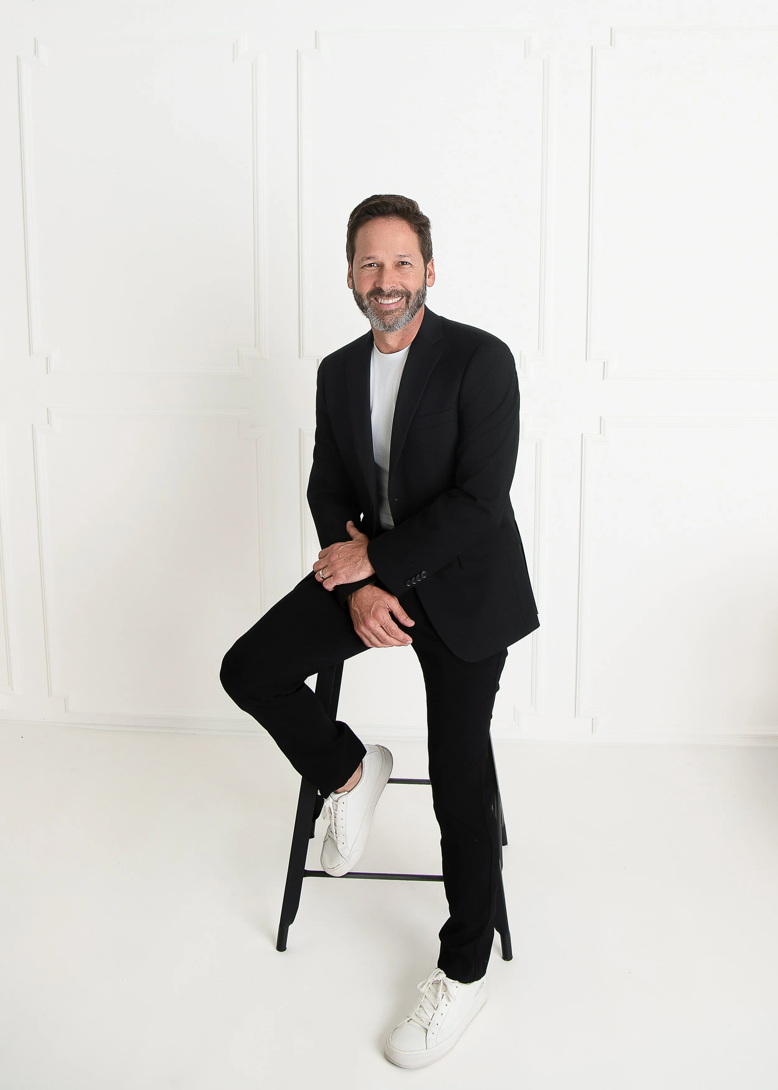 A middle-aged man with a beard smiling, wearing a black blazer, white shirt, black pants, white sneakers, sitting on a black stool against a white wall with decorative paneling.