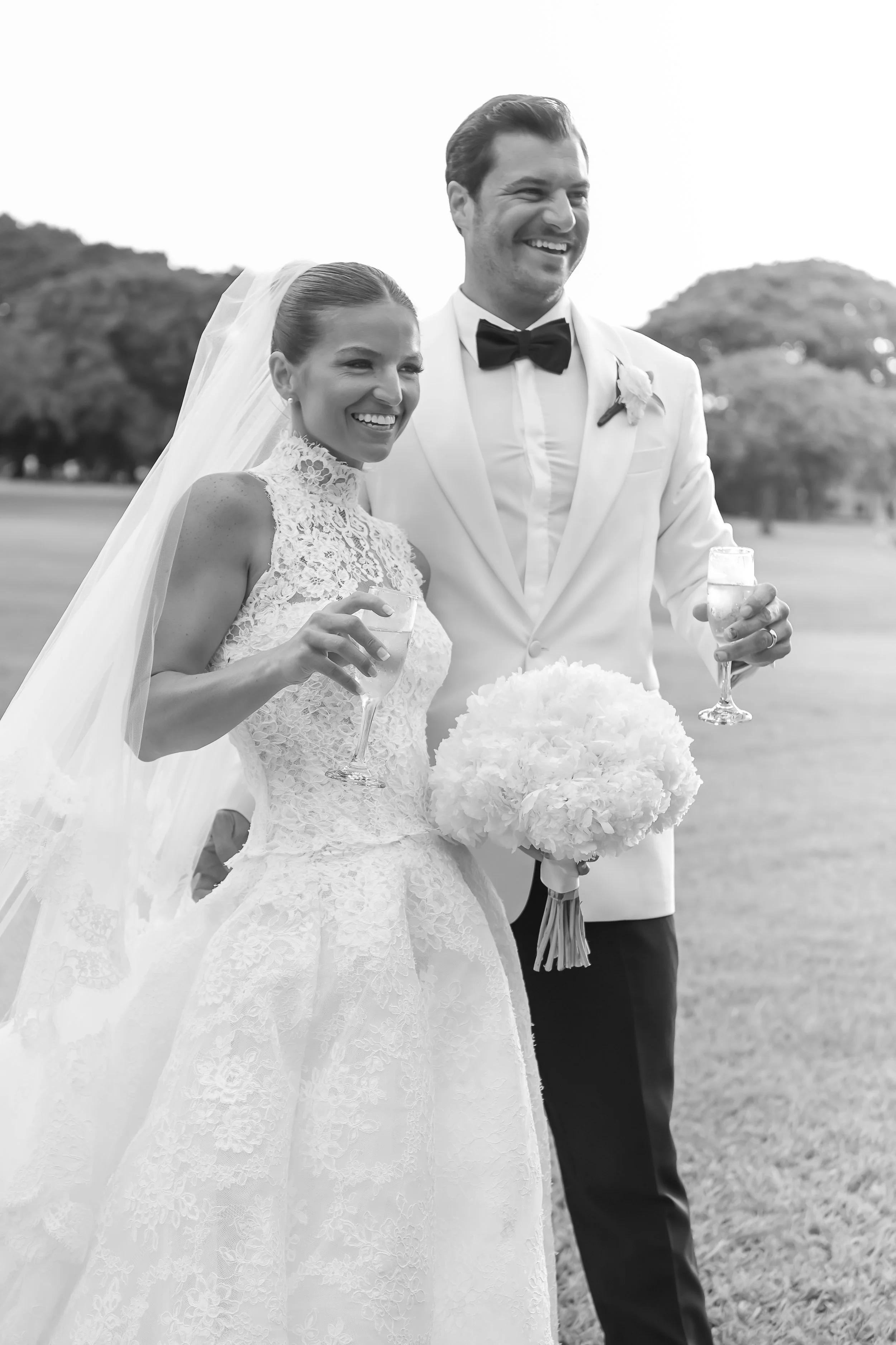 Black and white photo of a smiling bride and groom holding champagne glasses outdoors, with trees in the background.