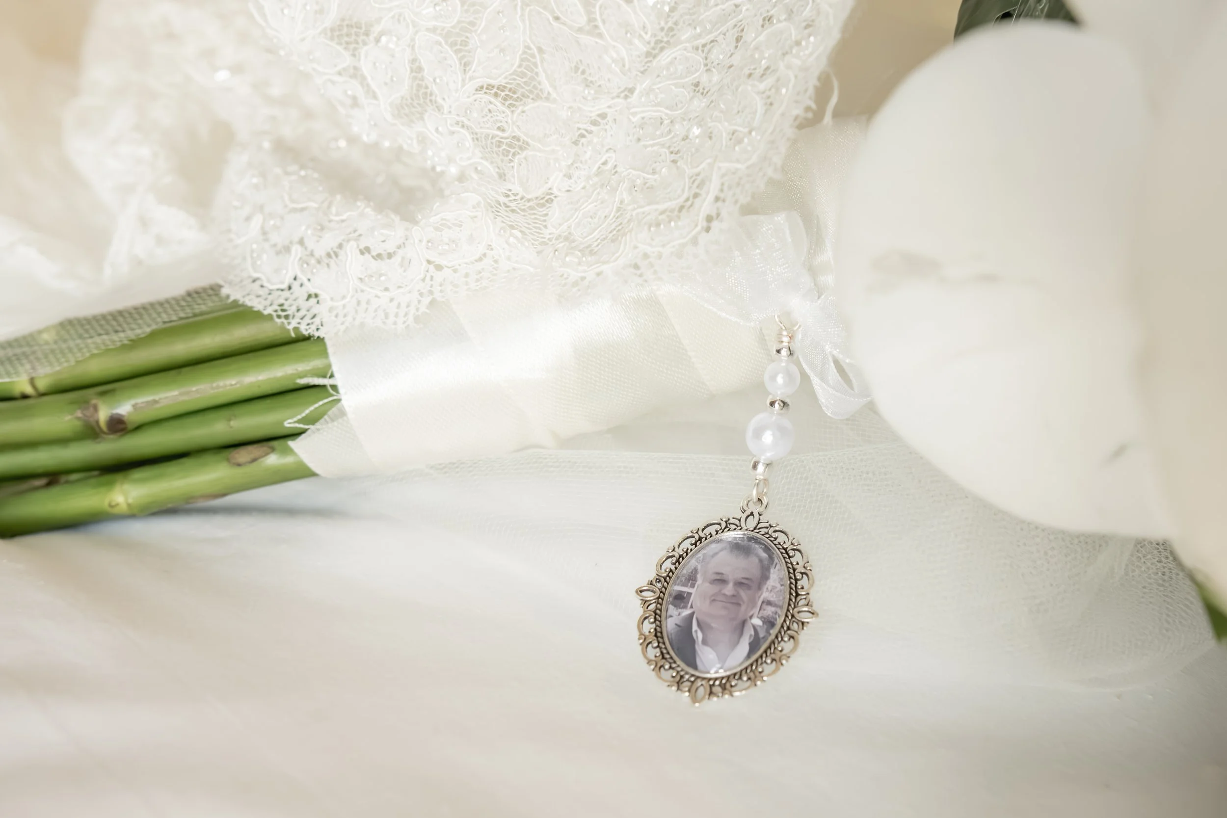 A close-up of a white lace wedding bouquet with fresh green bamboo stems underneath. Attached is a vintage photo pendant necklace with a black and white portrait of a smiling man.