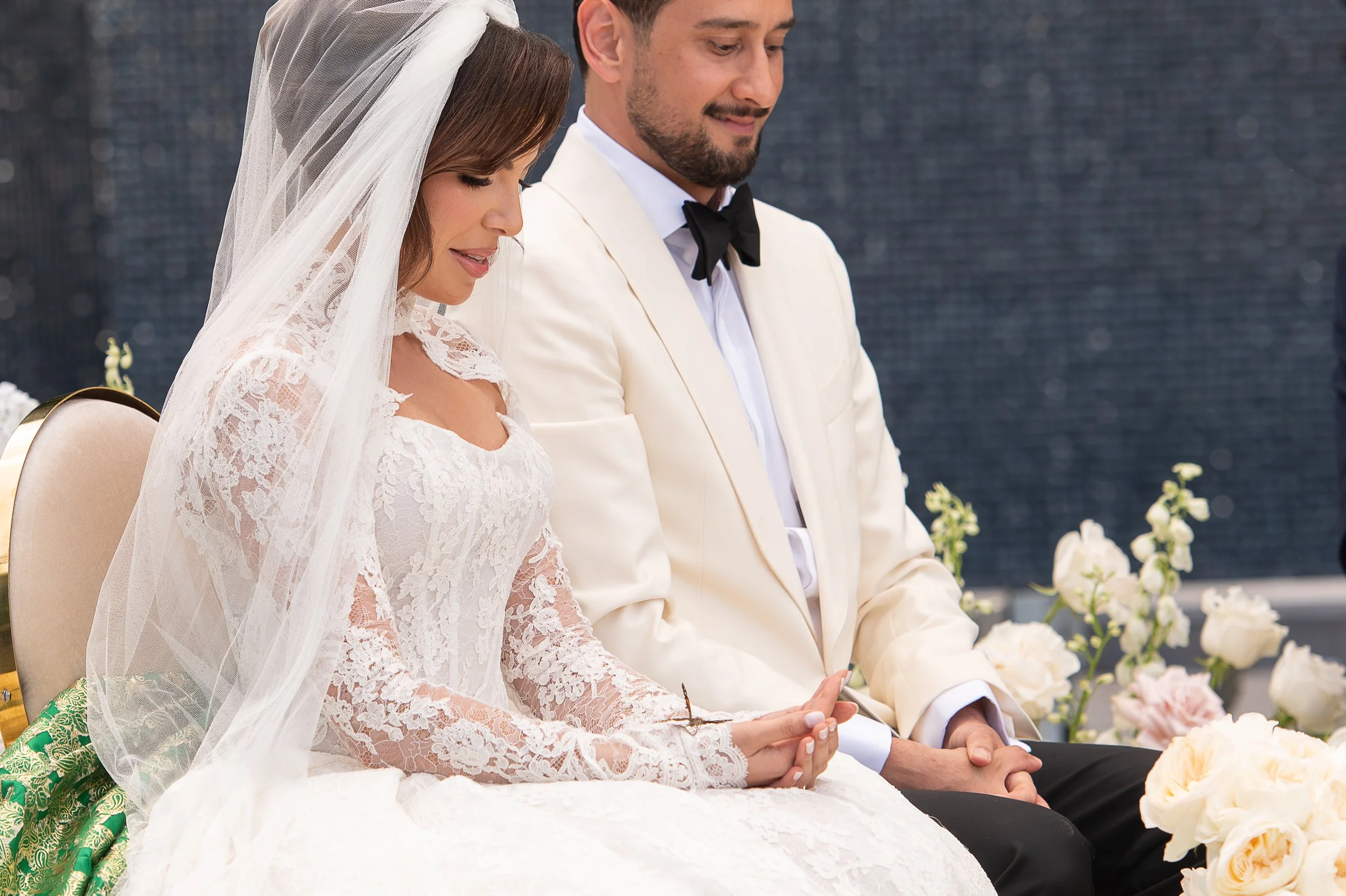 A bride and groom sitting side by side during their wedding ceremony, with the bride wearing a lace wedding dress and veil, and the groom in a white tuxedo with a black bow tie, surrounded by white flowers.