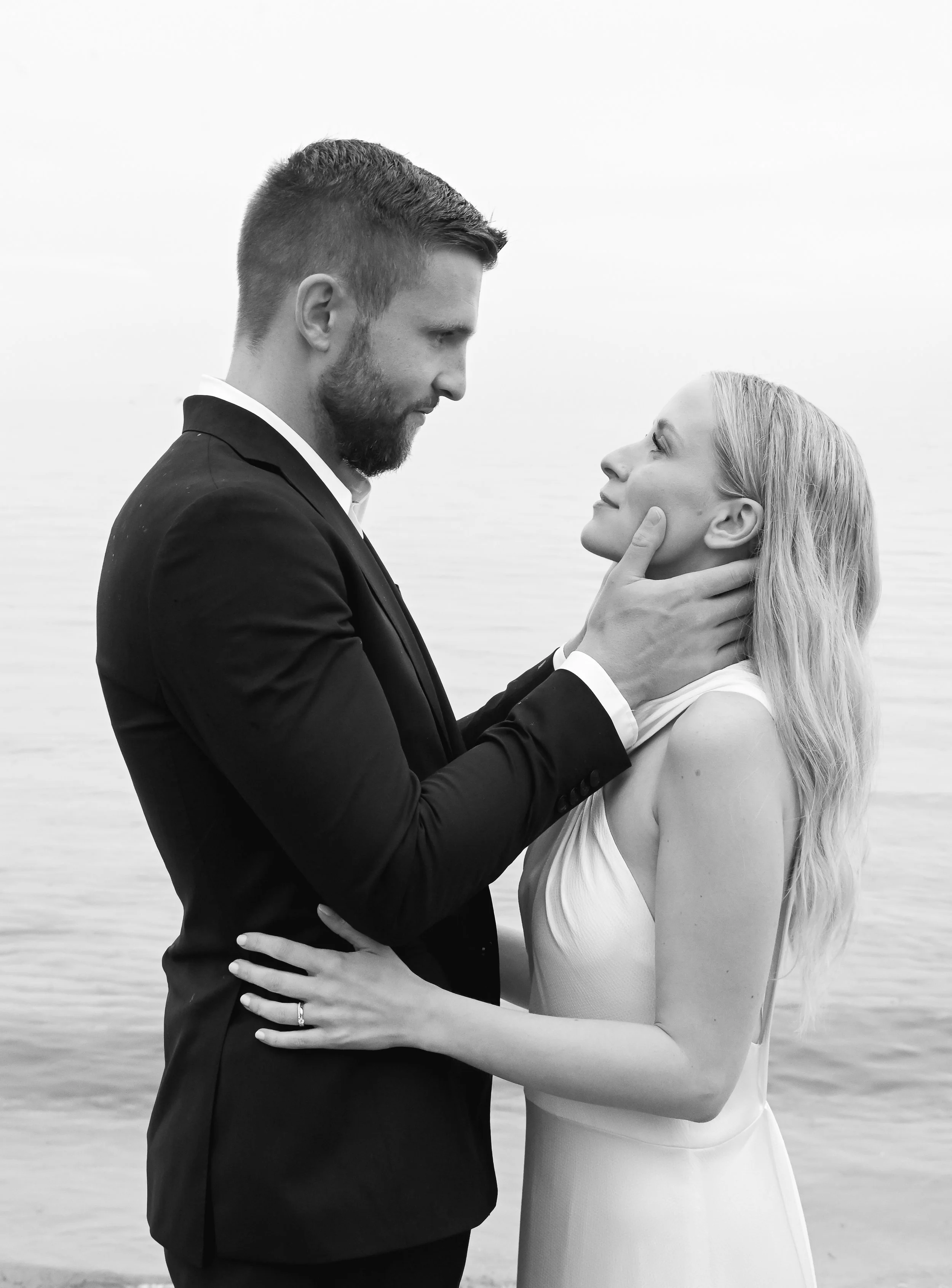 A black and white photo of a couple at the beach, the man in a suit holding the woman's face as they gaze into each other's eyes.
