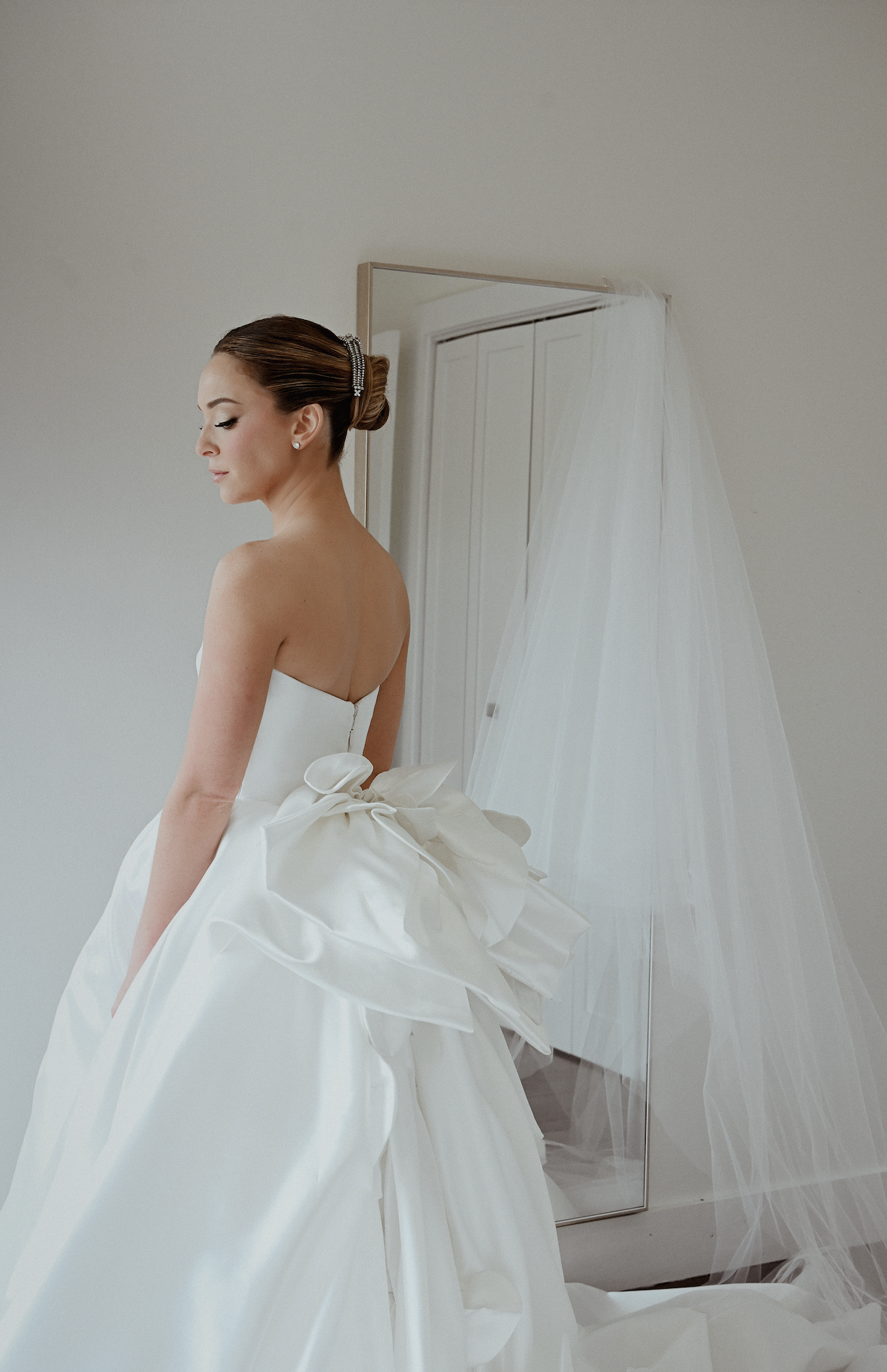 A bride in a wedding dress with large bow details, standing indoors near a mirror and a wedding veil.