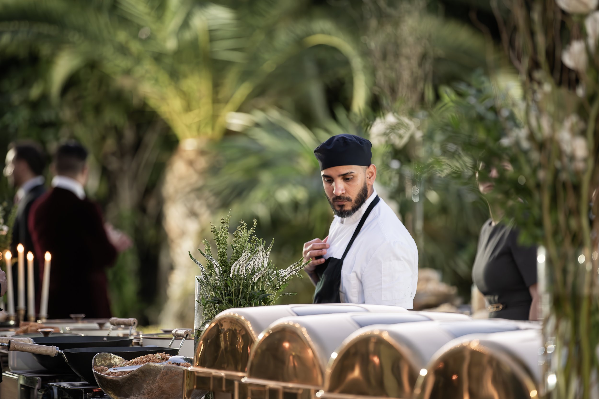 A chef in a black hat and white uniform stands behind a table with various dishes and decorative elements outdoors, surrounded by green foliage and trees.