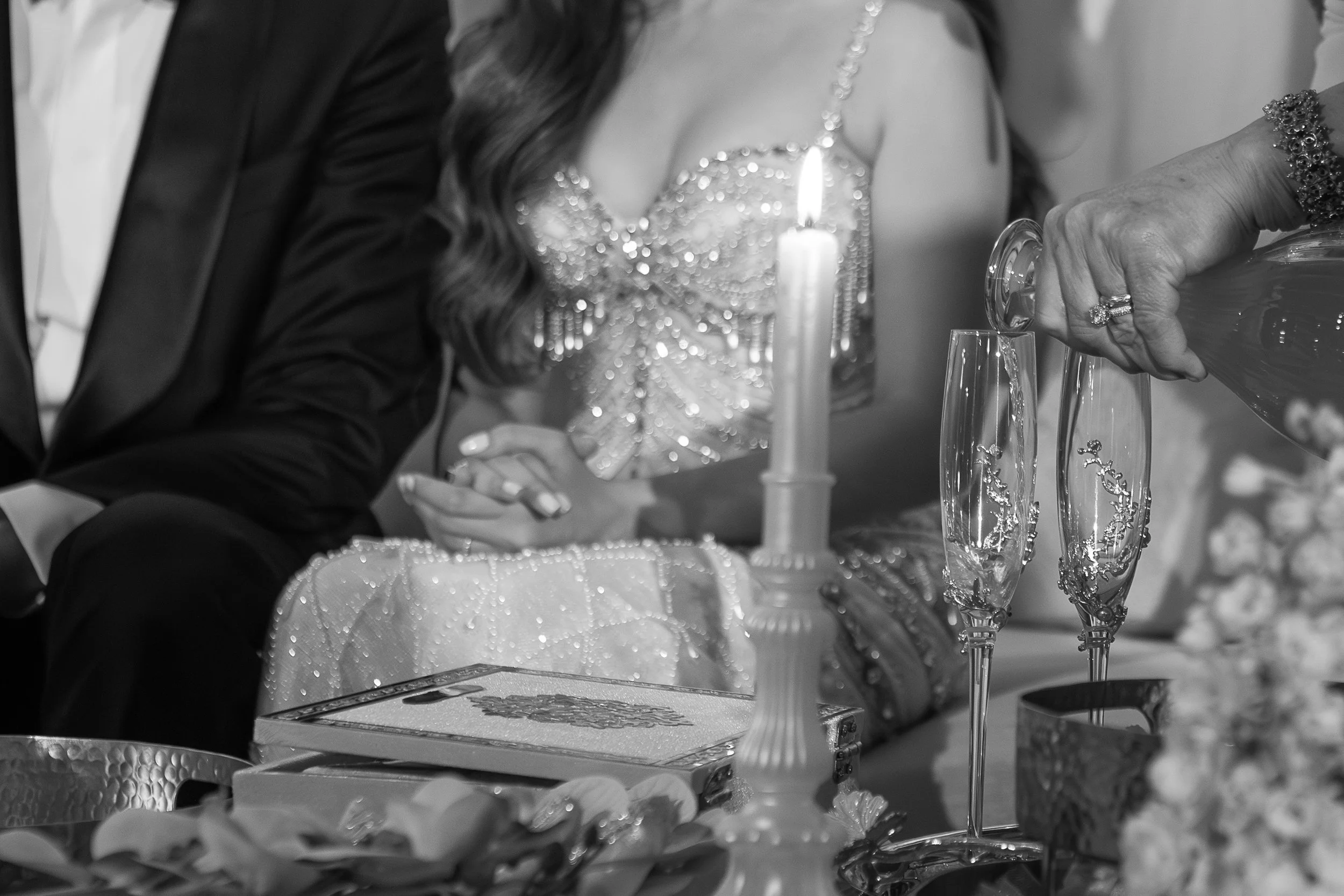 A black and white photo of a wedding scene where a couple is pouring champagne into glasses. The bride is wearing a bejeweled dress and jewelry, and the groom is in a tuxedo. There is a lit candle on the table and ornate glassware, indicating a celeb