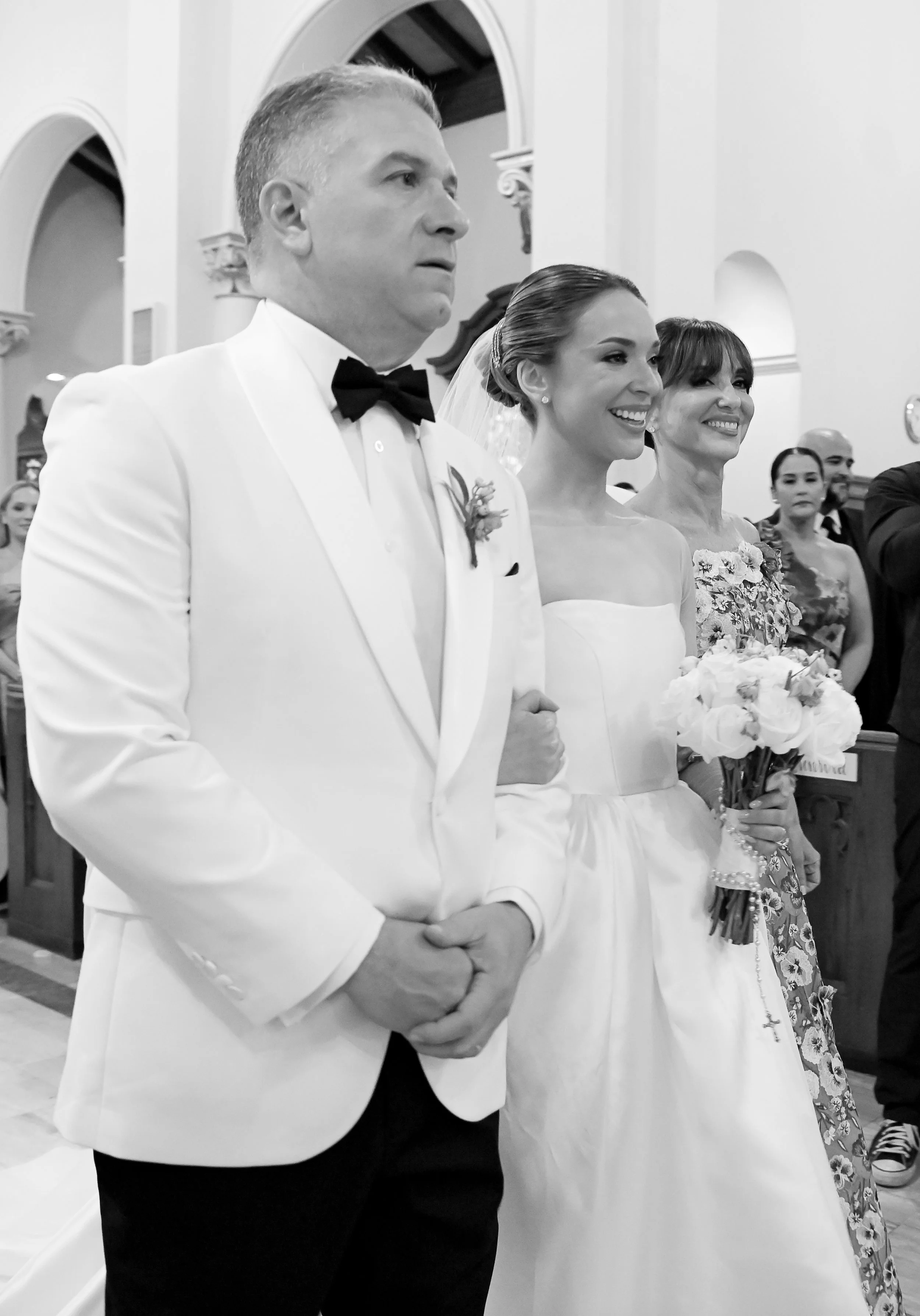 A black and white photo of a wedding ceremony inside a church, with the groom in a white tuxedo, black bow tie, and boutonniere, standing beside a smiling bride in a strapless white wedding dress holding a bouquet. There are guests in the background.