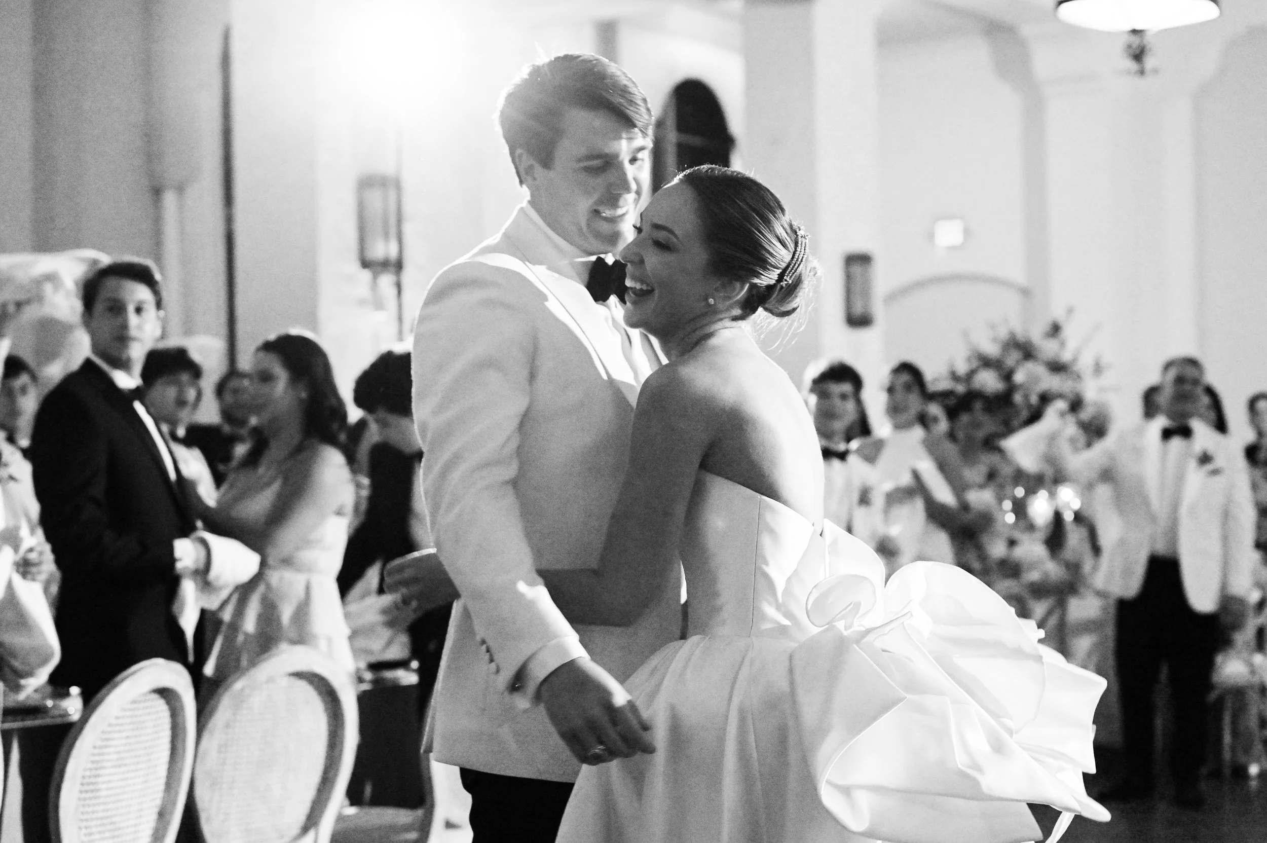 A couple in wedding attire dancing together at their wedding reception, surrounded by seated guests in a decorated indoor venue.