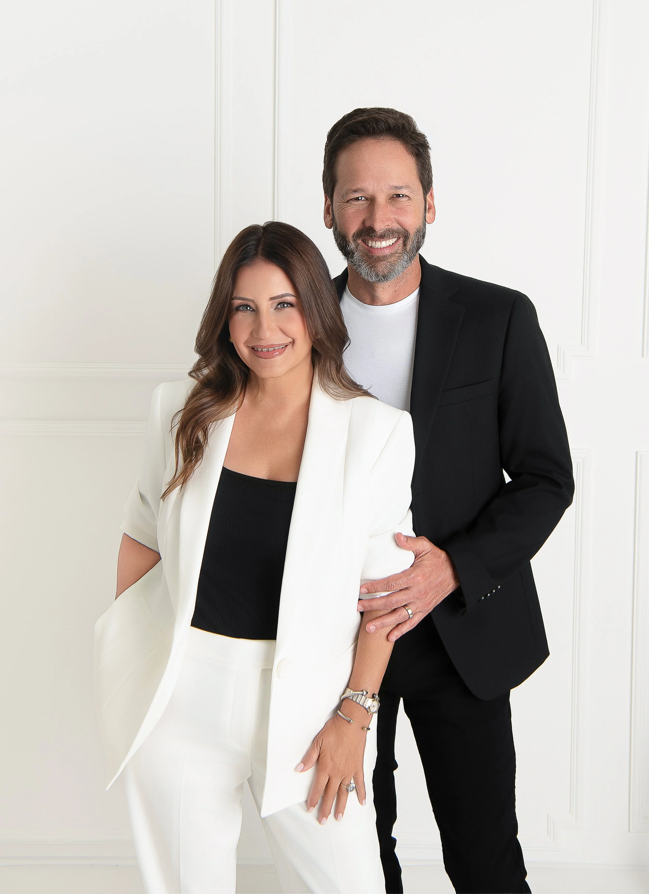 A man and woman dressed professionally, smiling while standing close to each other against a plain white background.