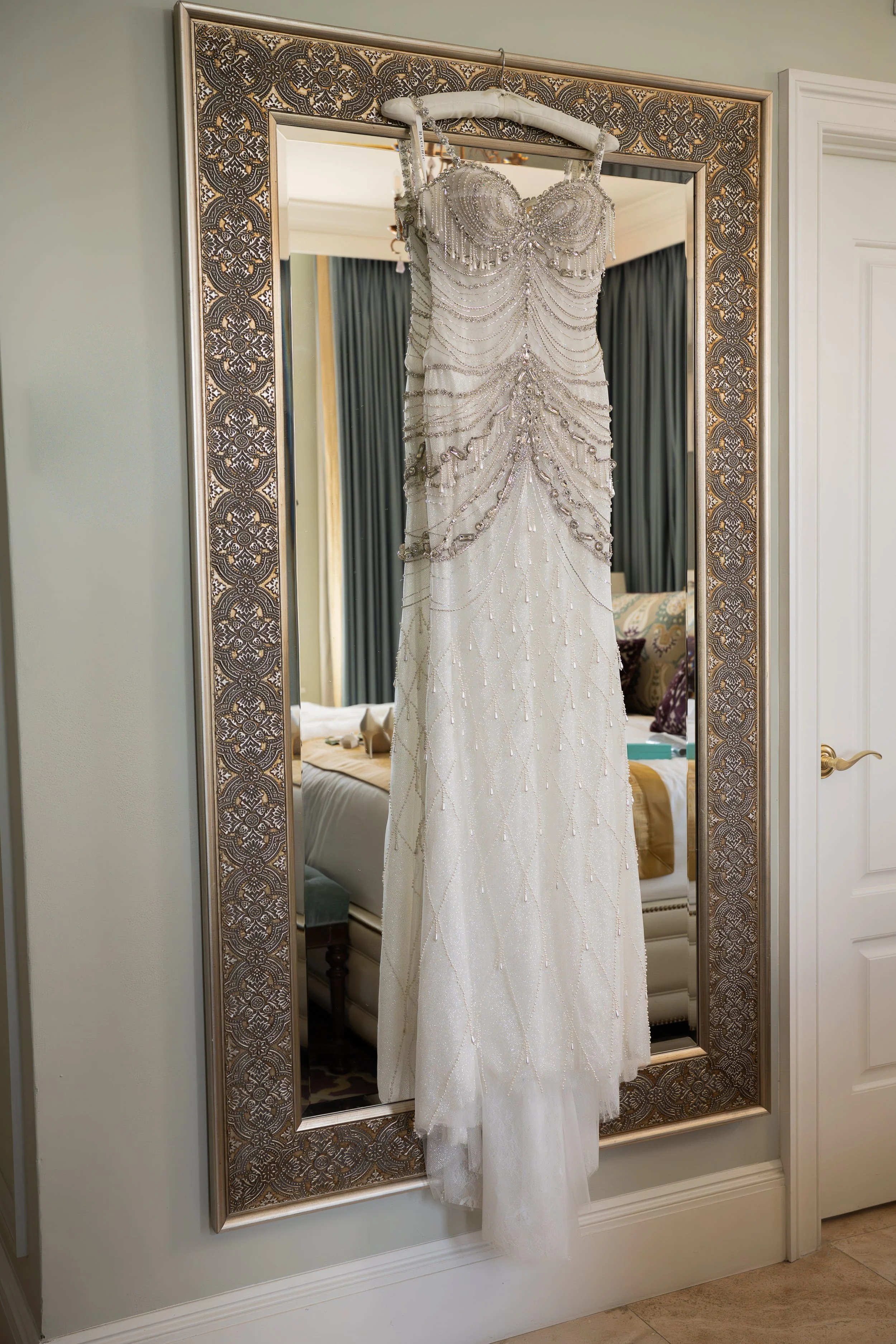 A wedding dress with intricate beading and ruching hangs on a padded hanger in front of a mirror with an ornate metal frame. The dress features a fitted bodice and a long flowing skirt, and is reflected in the mirror.