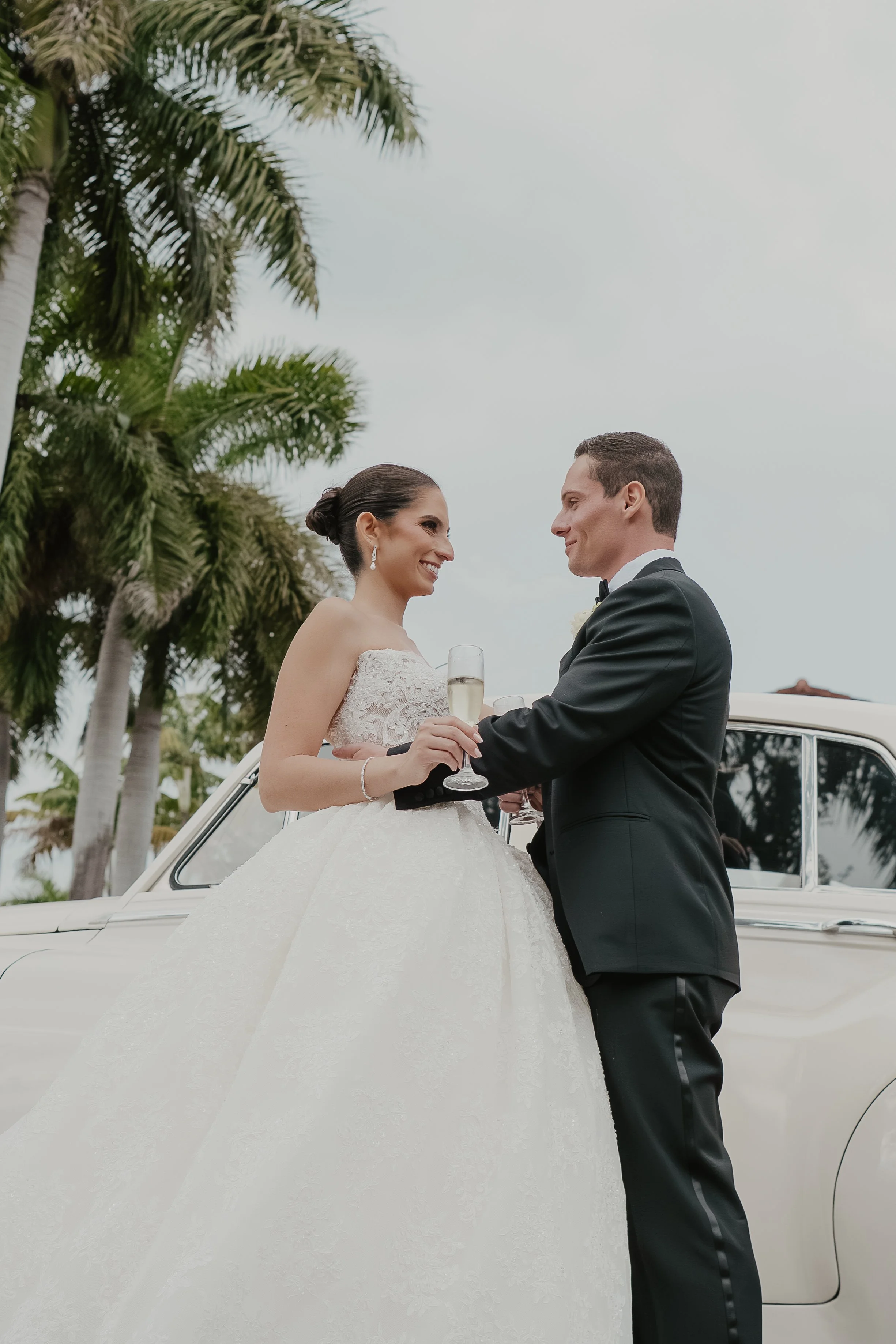 A bride and groom smiling and holding champagne glasses in front of a white vintage car, with palm trees and a cloudy sky in the background.
