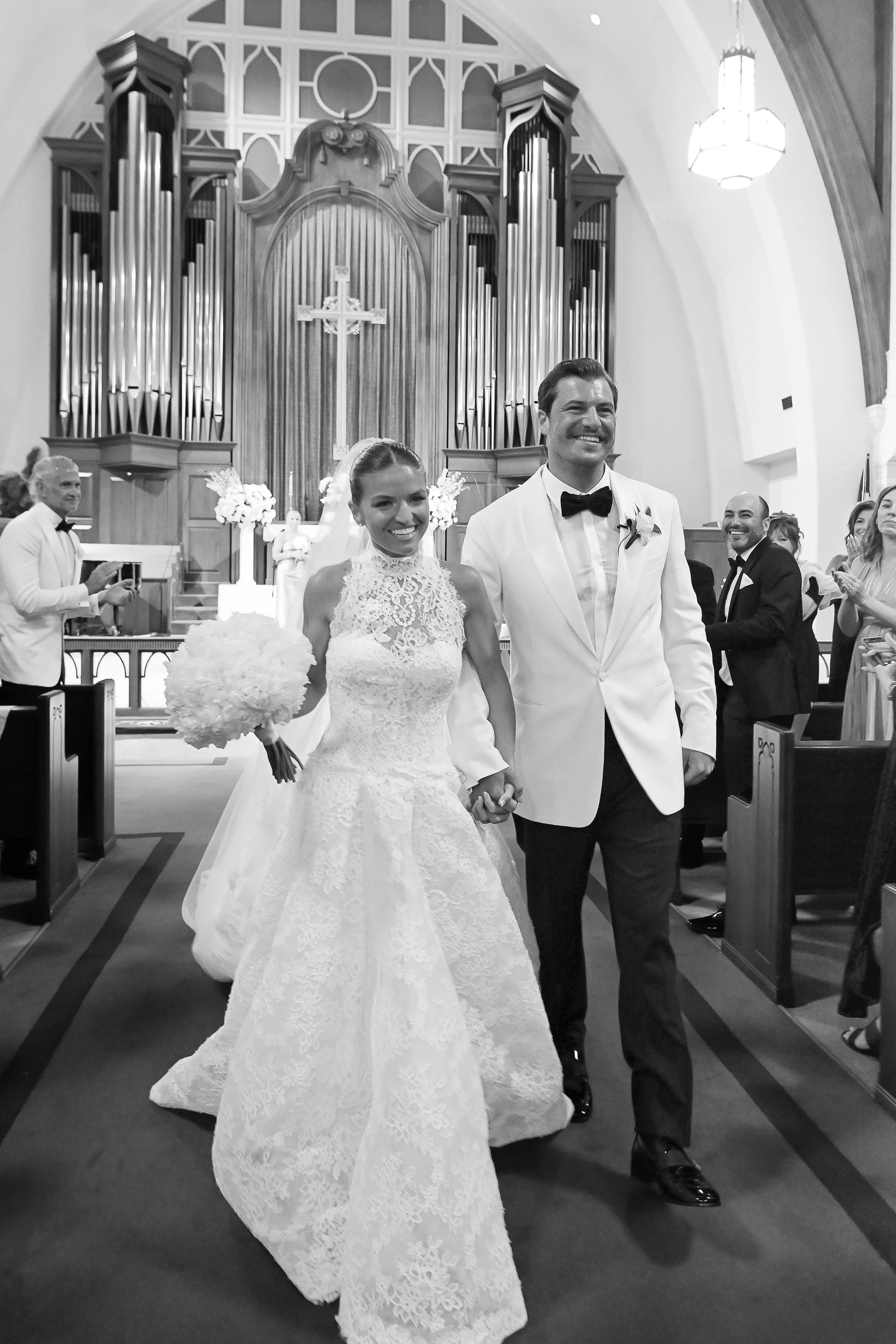 A bride and groom walking down the aisle of a church after getting married, with guests clapping and smiling in the background. The bride is holding a bouquet of flowers and wearing a lace wedding dress, while the groom is dressed in a white tuxedo w