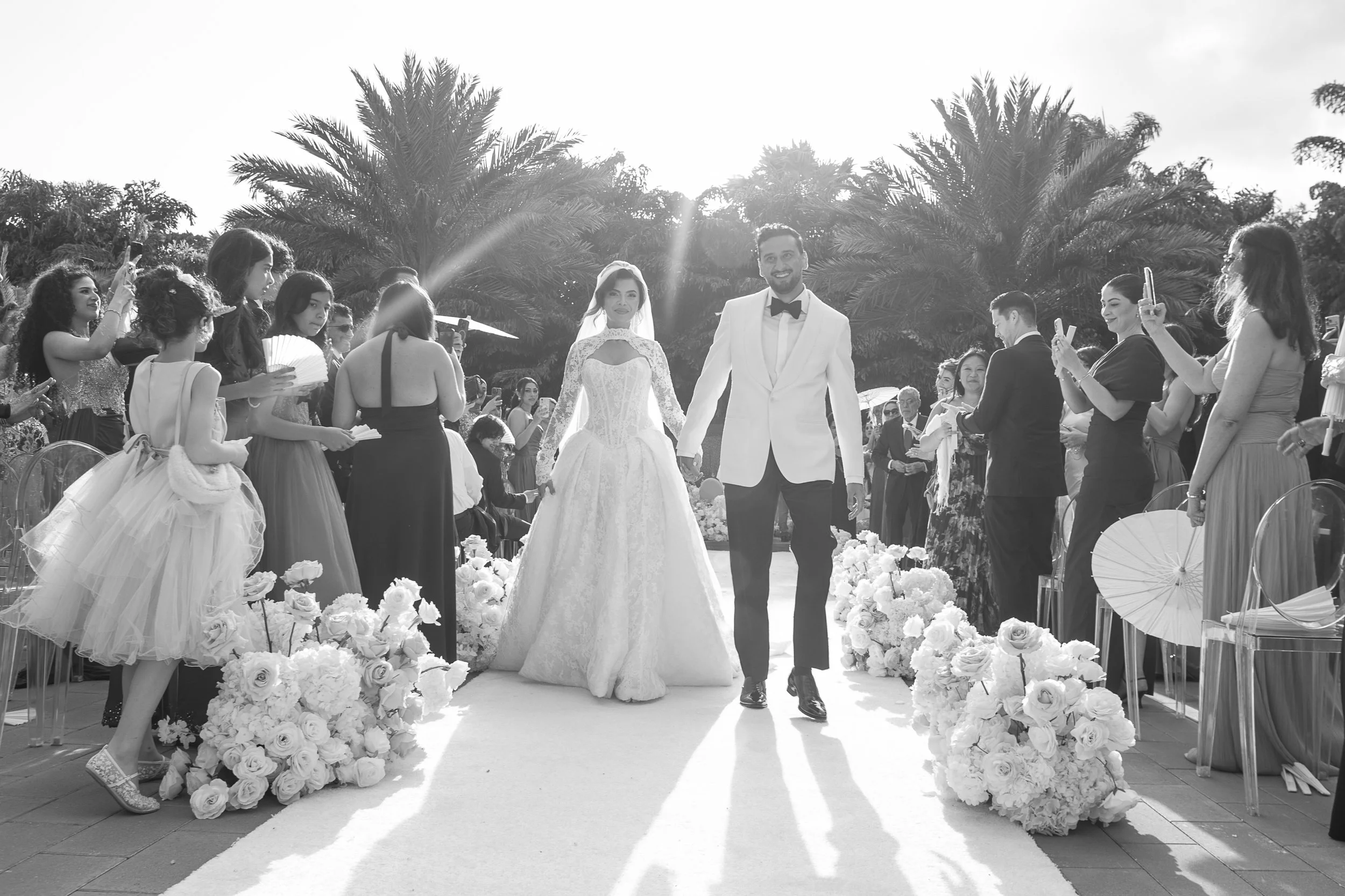 A black and white photo of a wedding scene with a bride and groom walking hand in hand down an aisle decorated with white roses, surrounded by guests taking photos, with a backdrop of palm trees and clear sky.