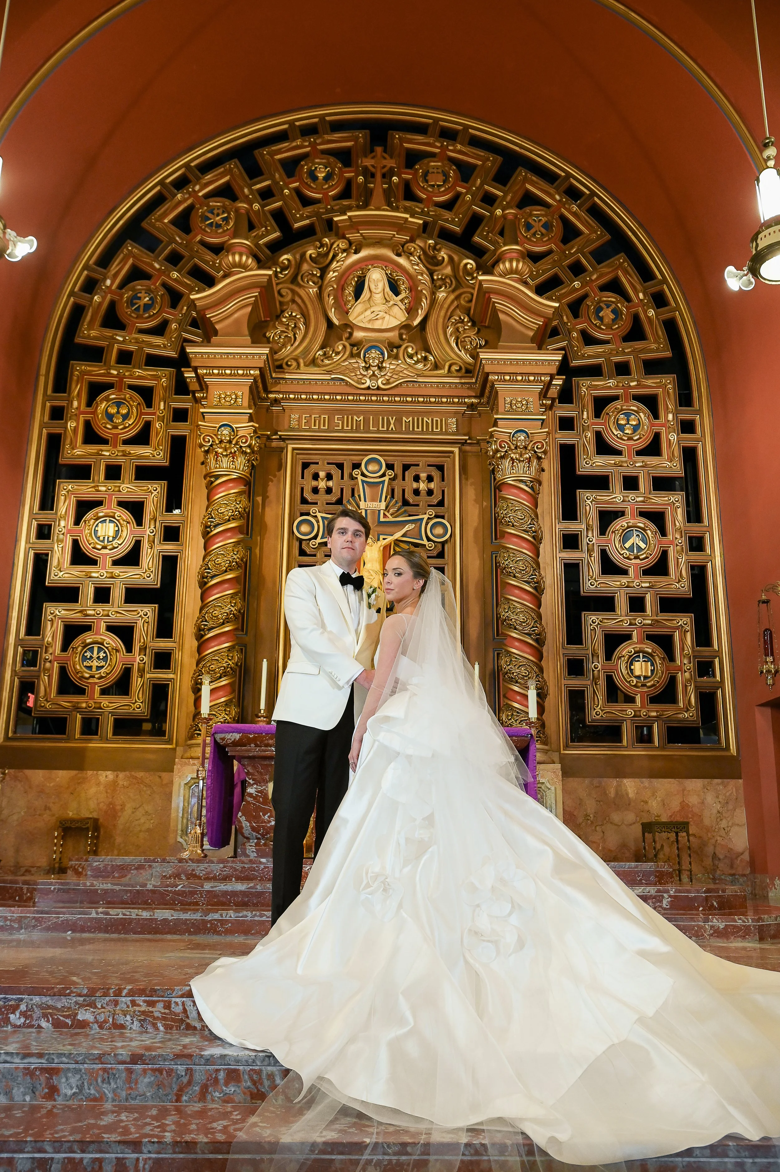 A bride and groom standing on the steps of a church altar, with ornate gold and wood decor behind them, during their wedding ceremony.