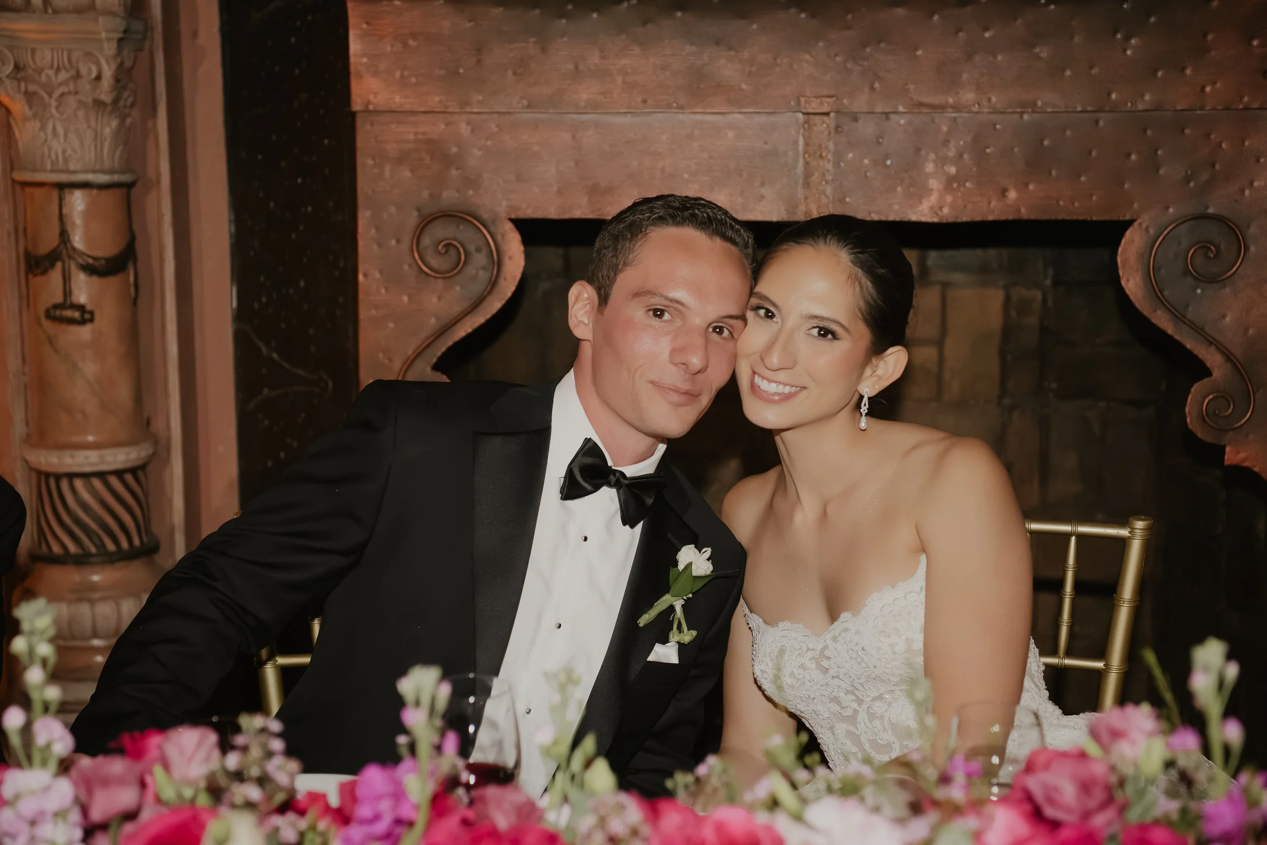 A newlywed couple sitting together at their wedding reception, smiling for the camera. The groom is wearing a black tuxedo with a bow tie, and the bride is in a white lace wedding dress with earrings. They are seated behind a table decorated with pin
