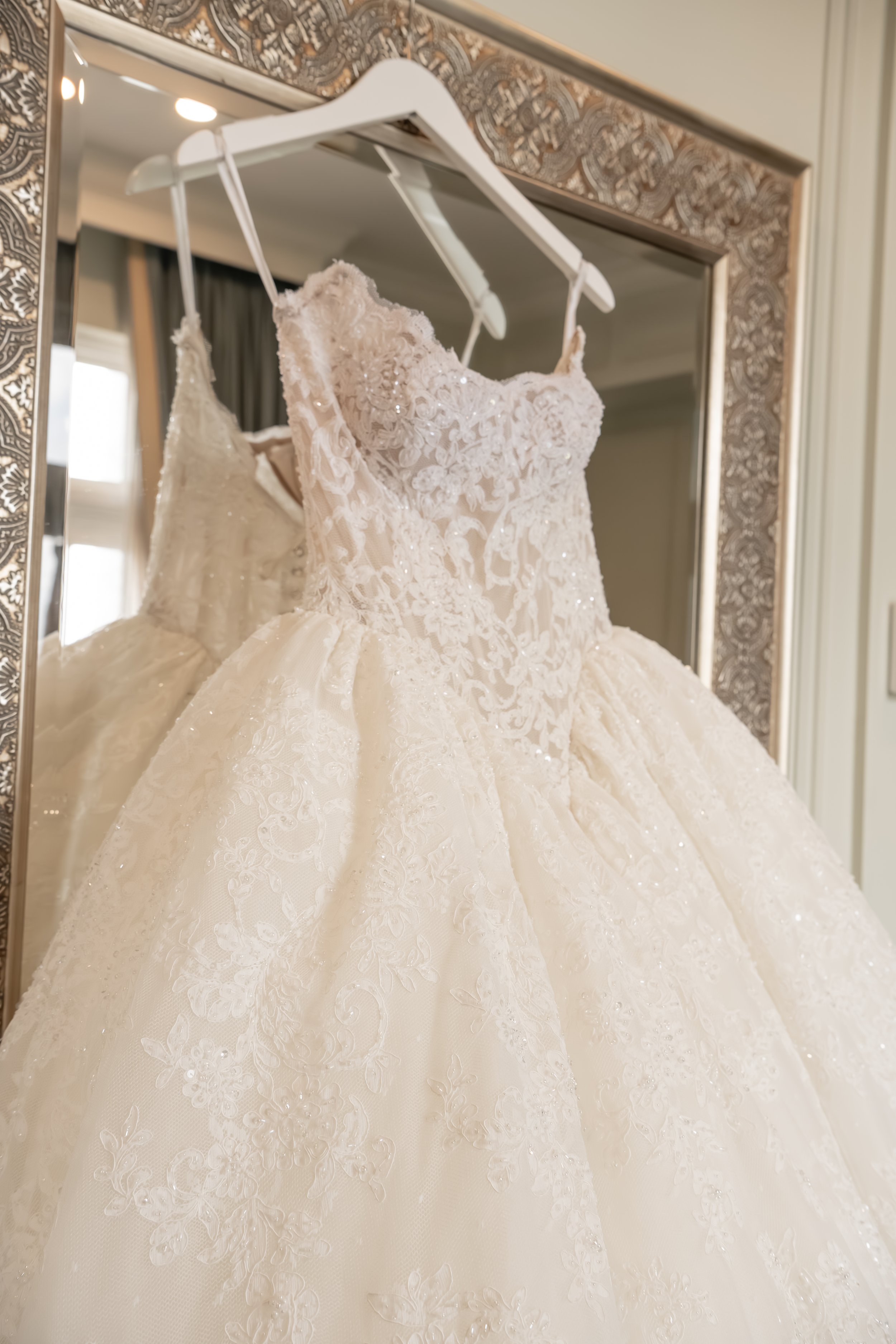 A wedding dress with lace details hanging on a white hanger, displayed in front of a mirror with an ornate framed border.