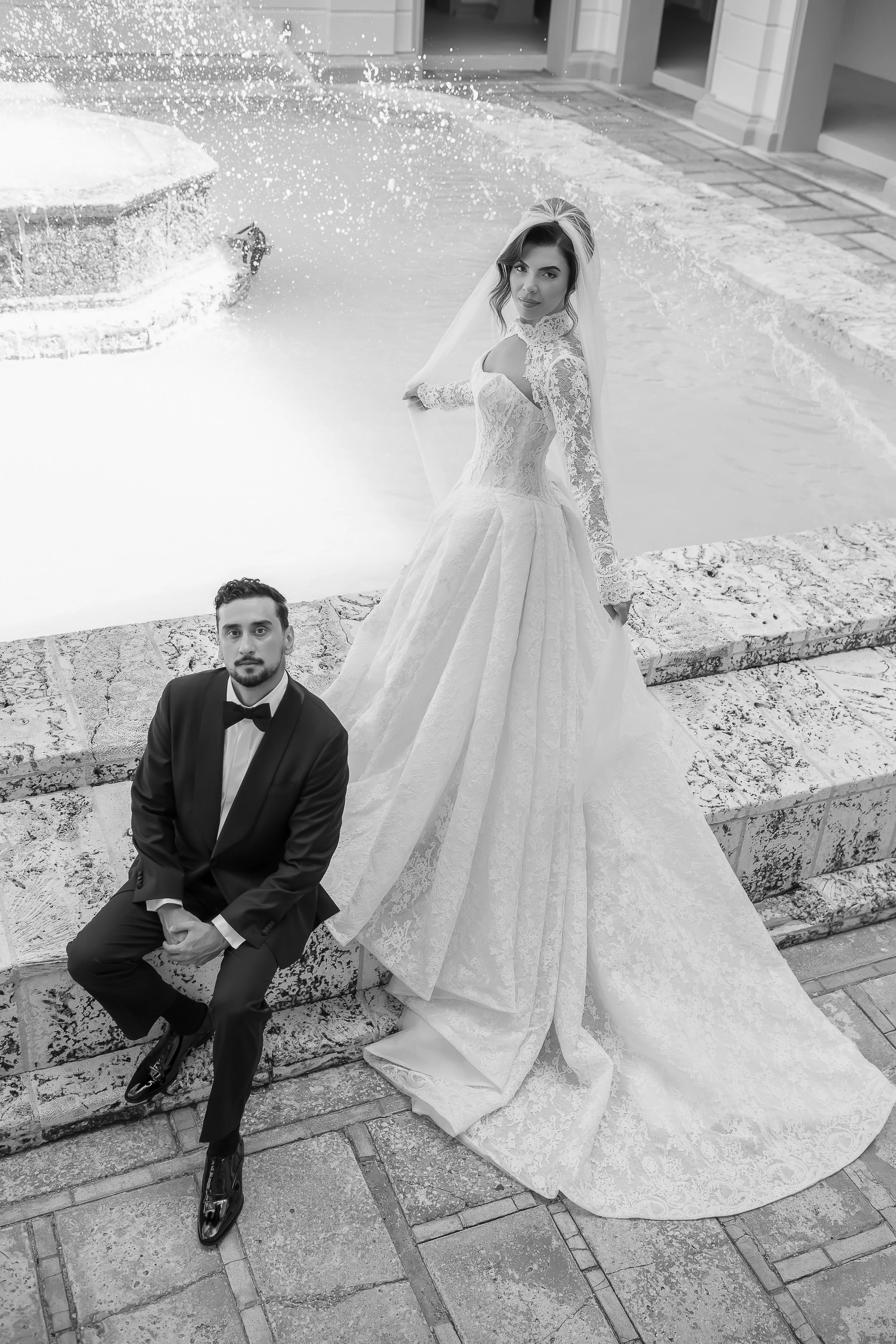 A bride in a wedding dress standing by a pool with an umbrella in the background, and a groom sitting on a stone wall next to her, outdoors near a building.