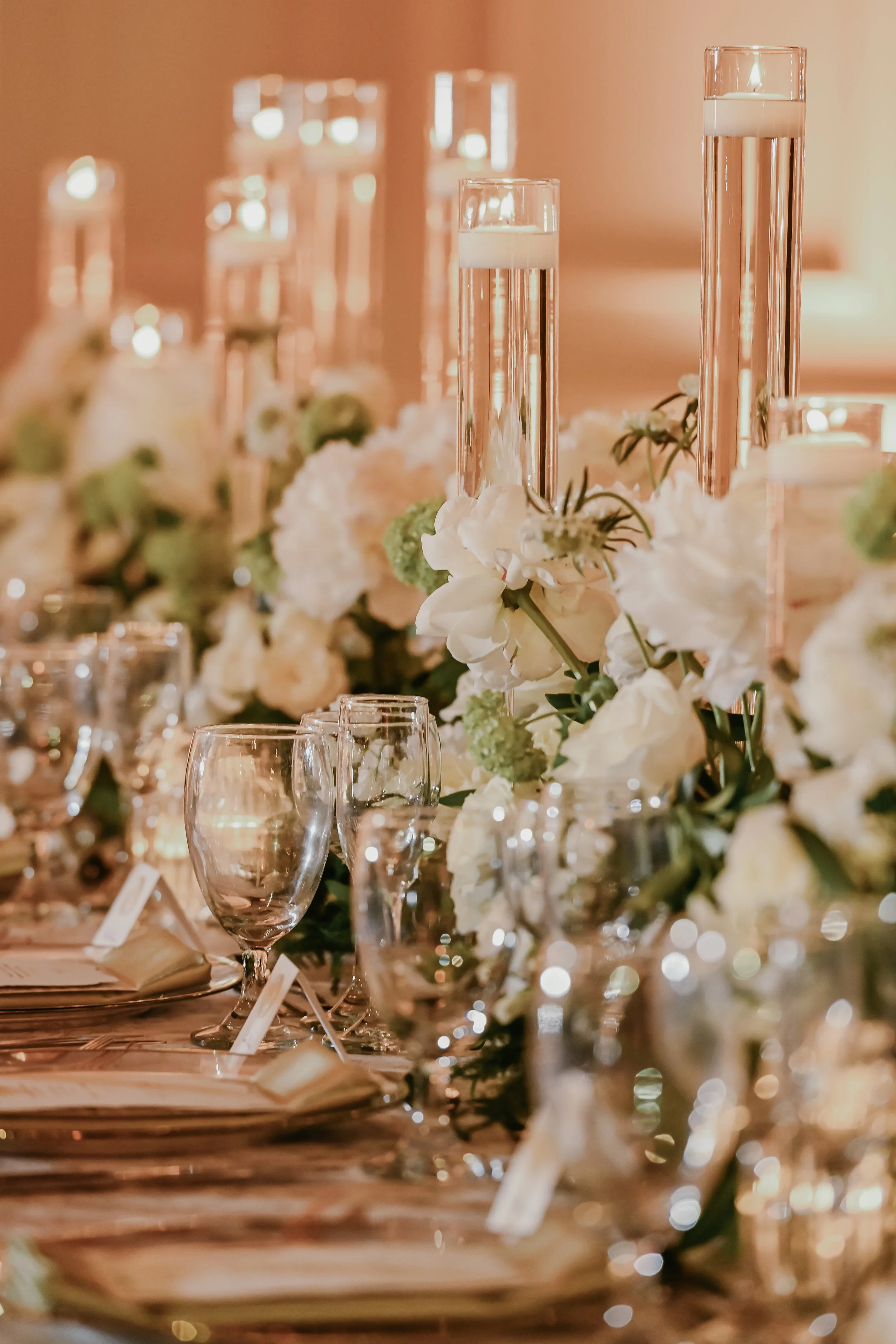 Elegant table setting with tall glass candleholders, white and green floral arrangements, wine glasses, and dinner plates, suitable for a wedding or formal event, decorated with soft warm lighting.