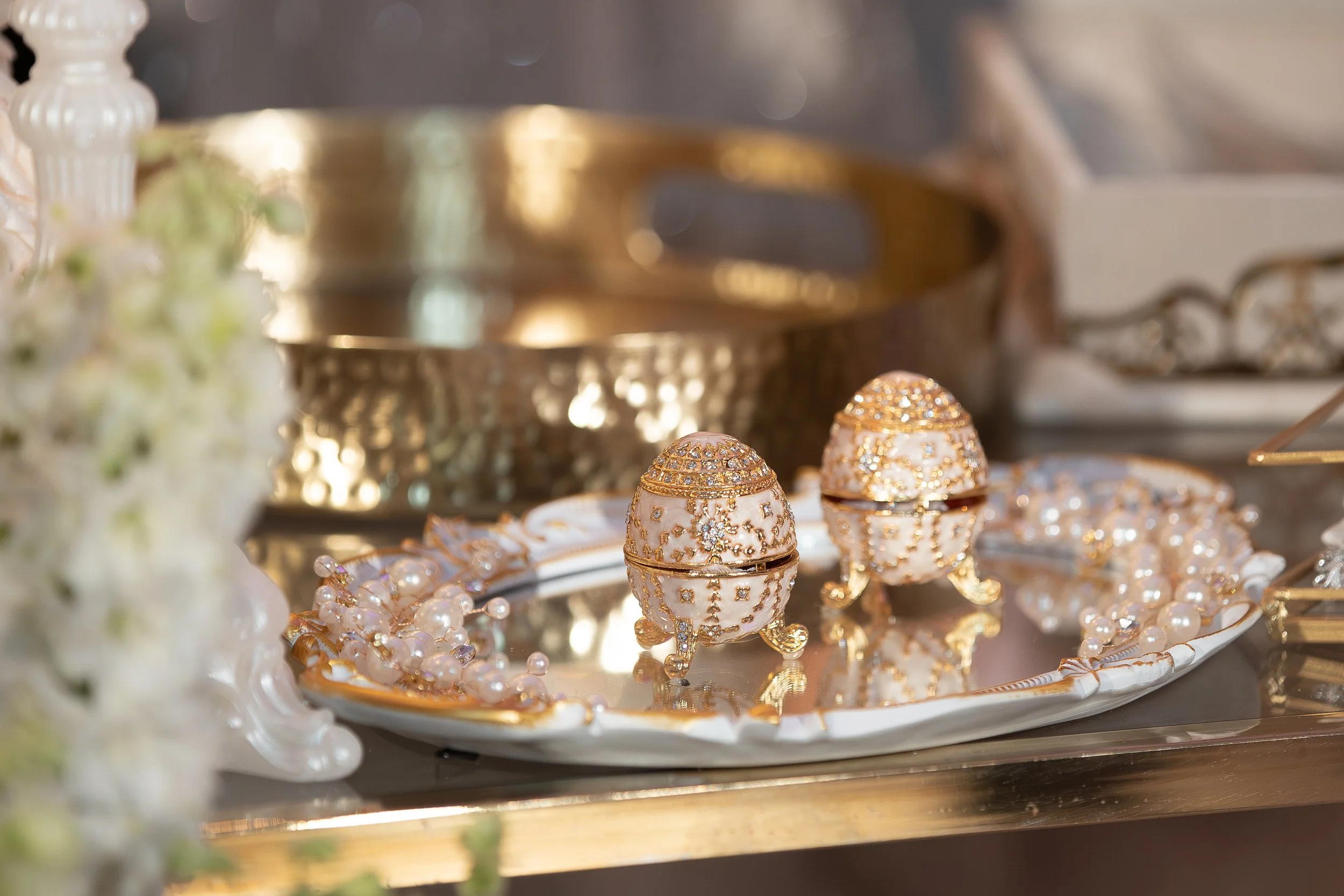 A tray of white and gold decorative jewelry, including rings, earrings, and a necklace, with intricate designs and embedded rhinestones, on a shiny surface with a blurred background.