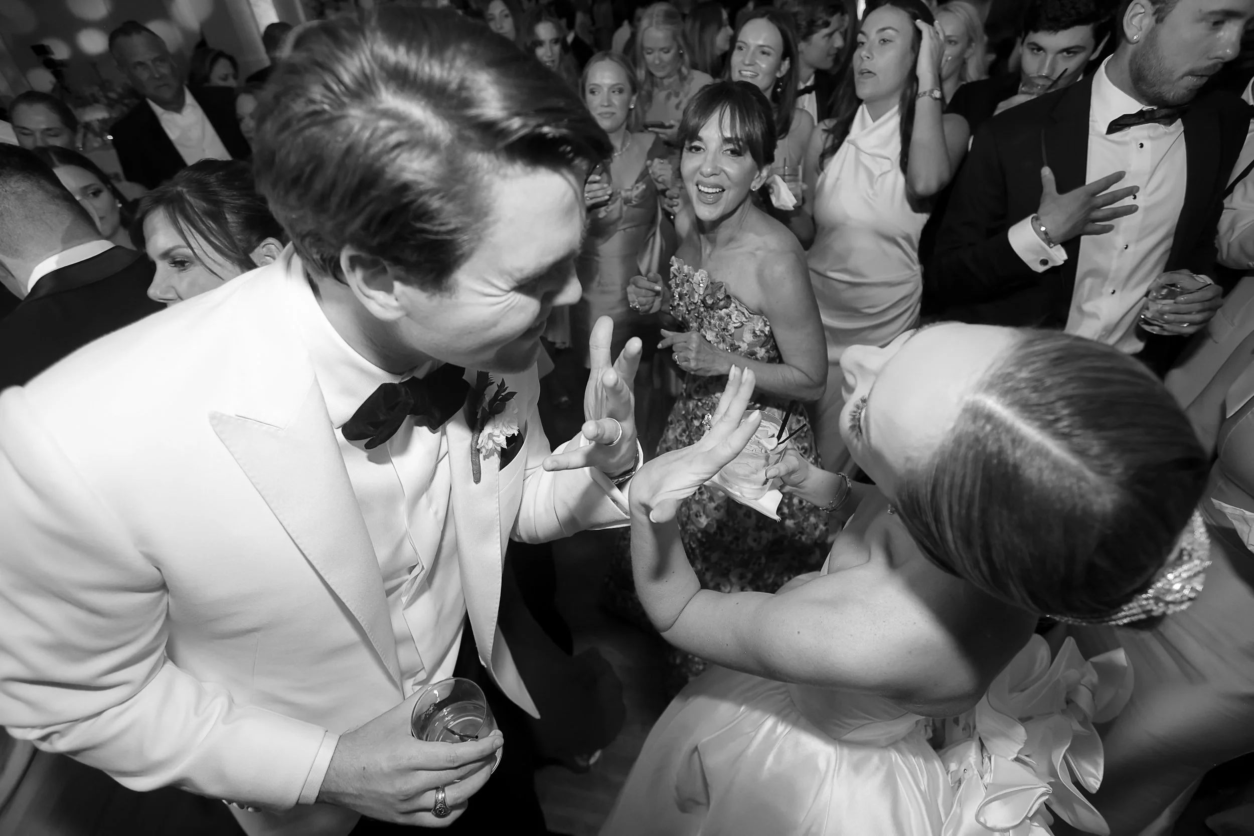 A black and white photo of a crowded dance floor at a formal event. People are dressed in tuxedos and elegant dresses, engaging in conversation and dancing.