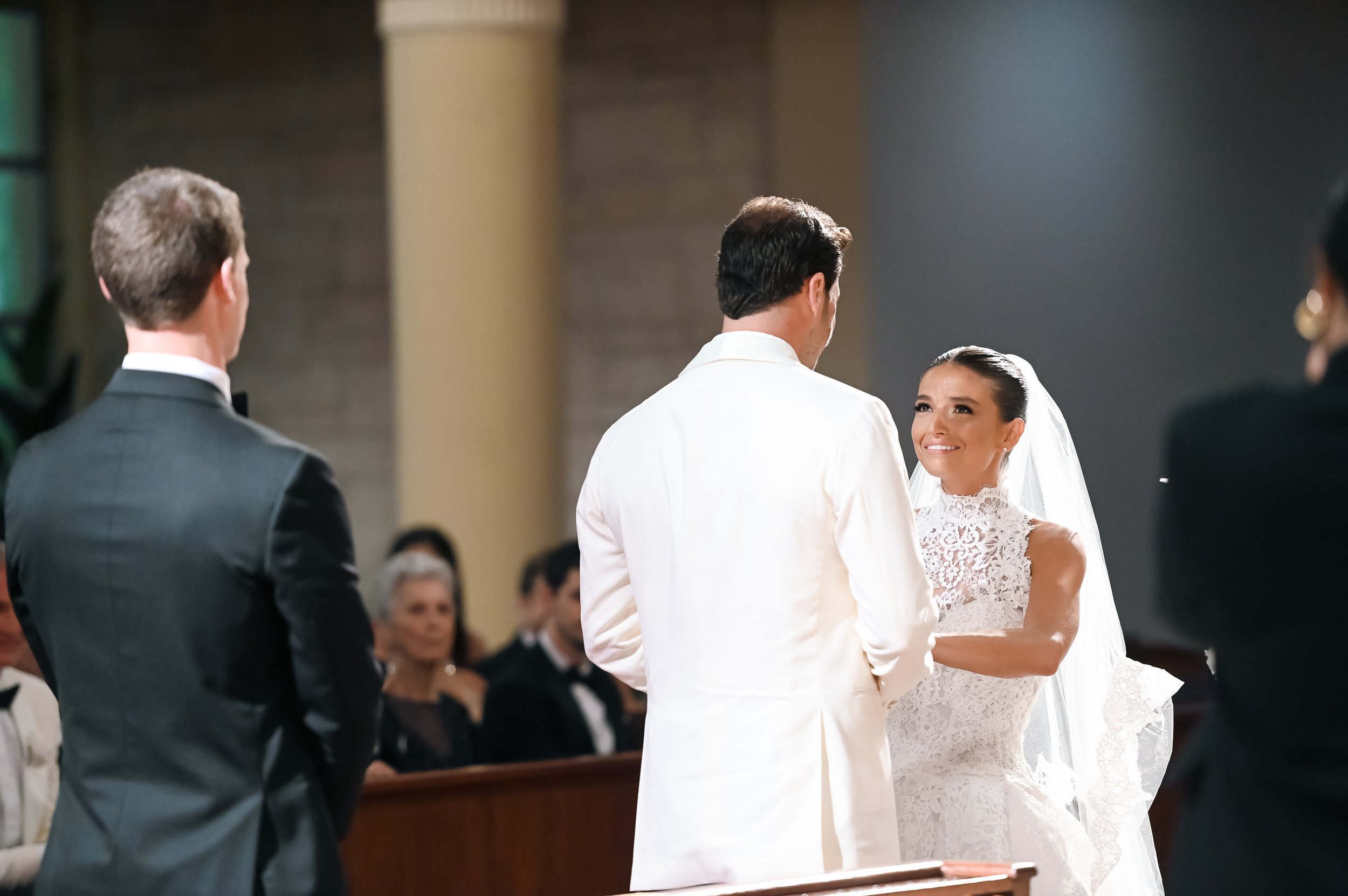 A bride and groom exchange vows during a wedding ceremony, with a priest or officiant standing between them. The bride is smiling and wearing a lace dress with a veil, while the groom is in a white suit jacket. Other guests are seated in the backgrou