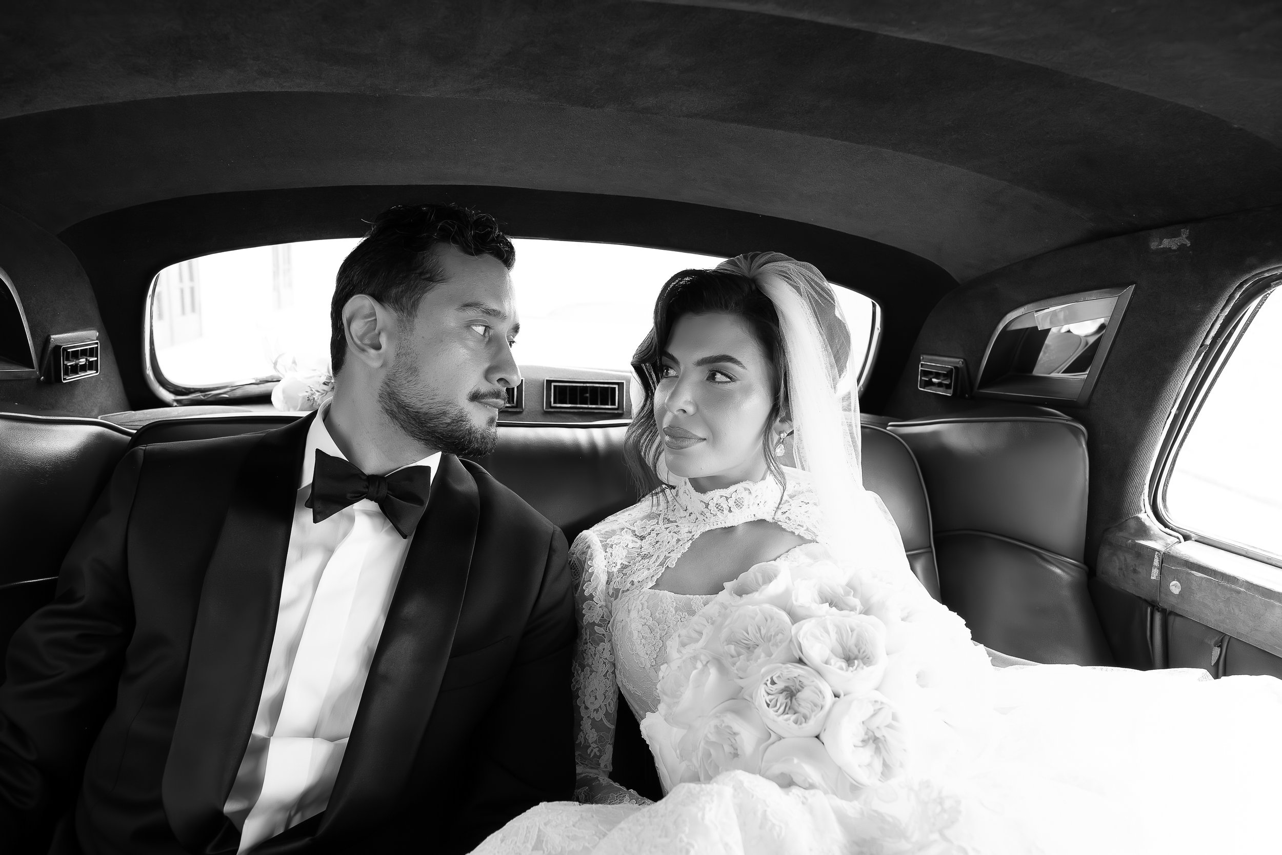 A black and white photo of a bride and groom inside a limousine, looking at each other. The groom is wearing a tuxedo with a bowtie, and the bride is in a lace wedding dress holding a bouquet of roses. They are sitting close together in the backseat.