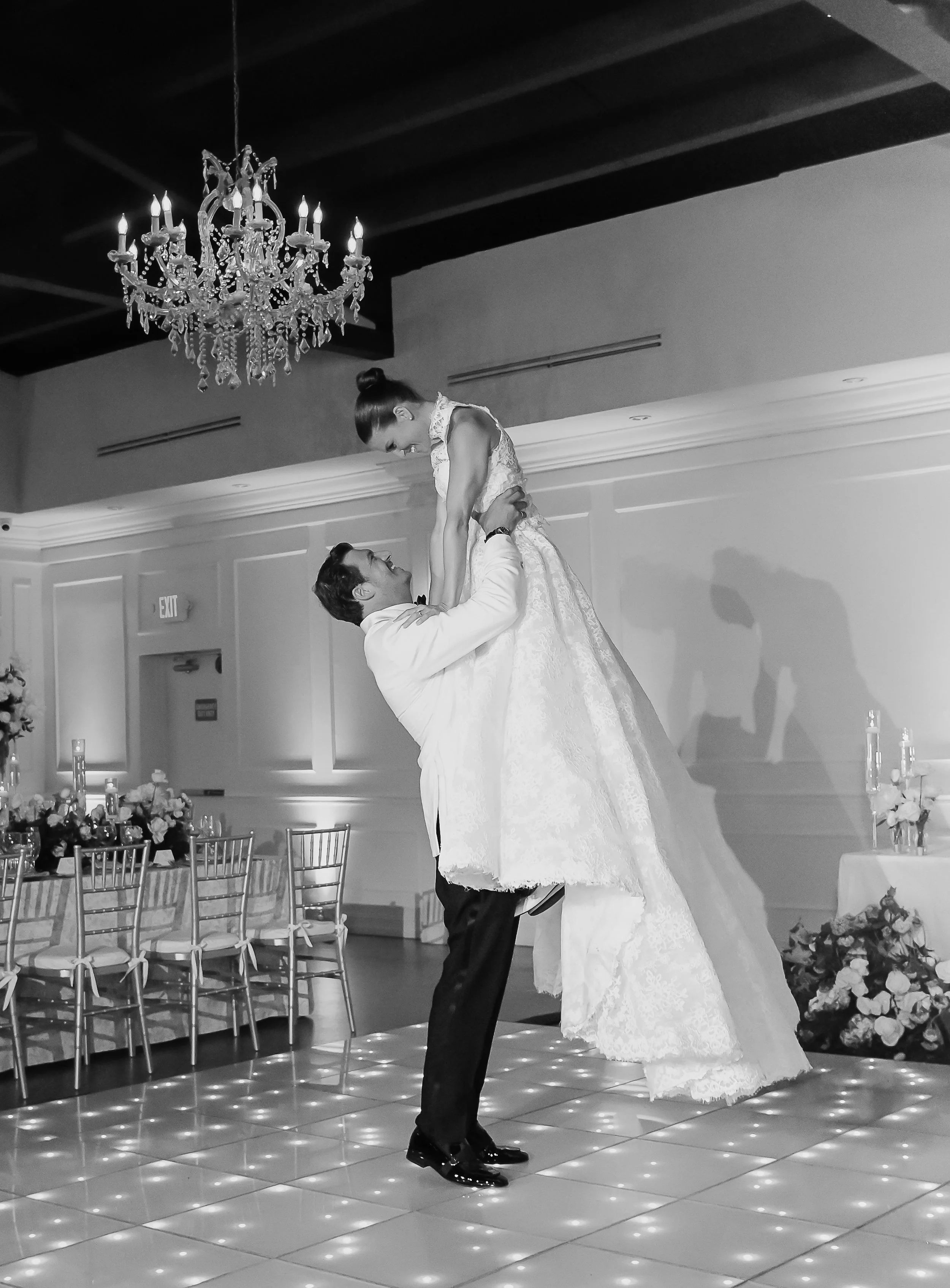 A black and white photo of a wedding reception with a chandelier hanging from the ceiling. A groom is lifting and dancing with the bride, who is wearing a gown. They are on a dance floor with a tiled pattern, and there are tables decorated with flowe