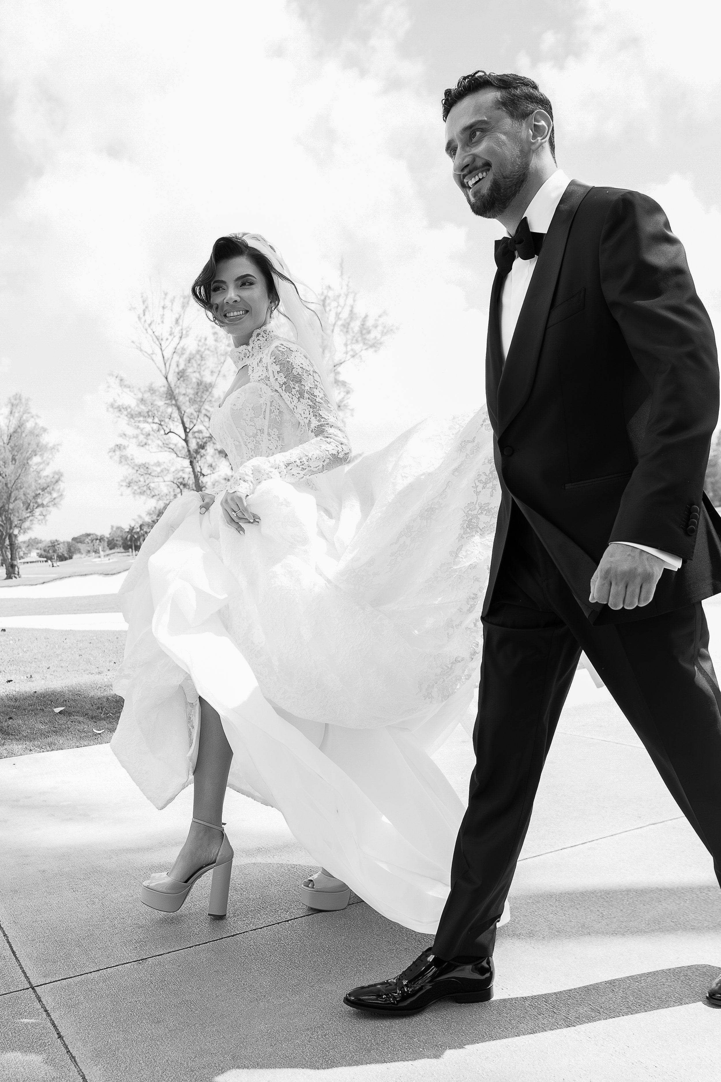 A bride and groom in formal wedding attire walking outdoors, with the bride lifting her dress to show high heels and the groom smiling.