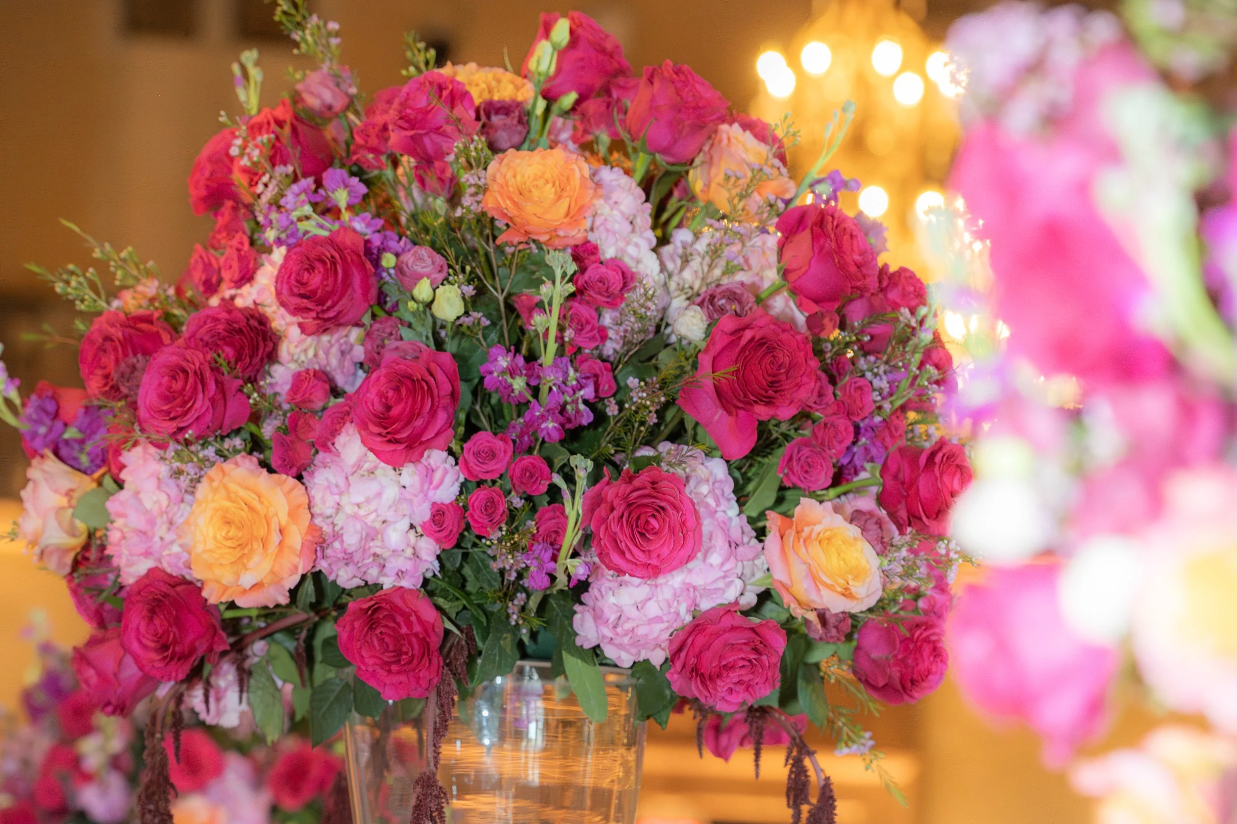 A vibrant bouquet of pink, orange, and purple flowers in a glass vase on a table with blurred warm lighting in the background.