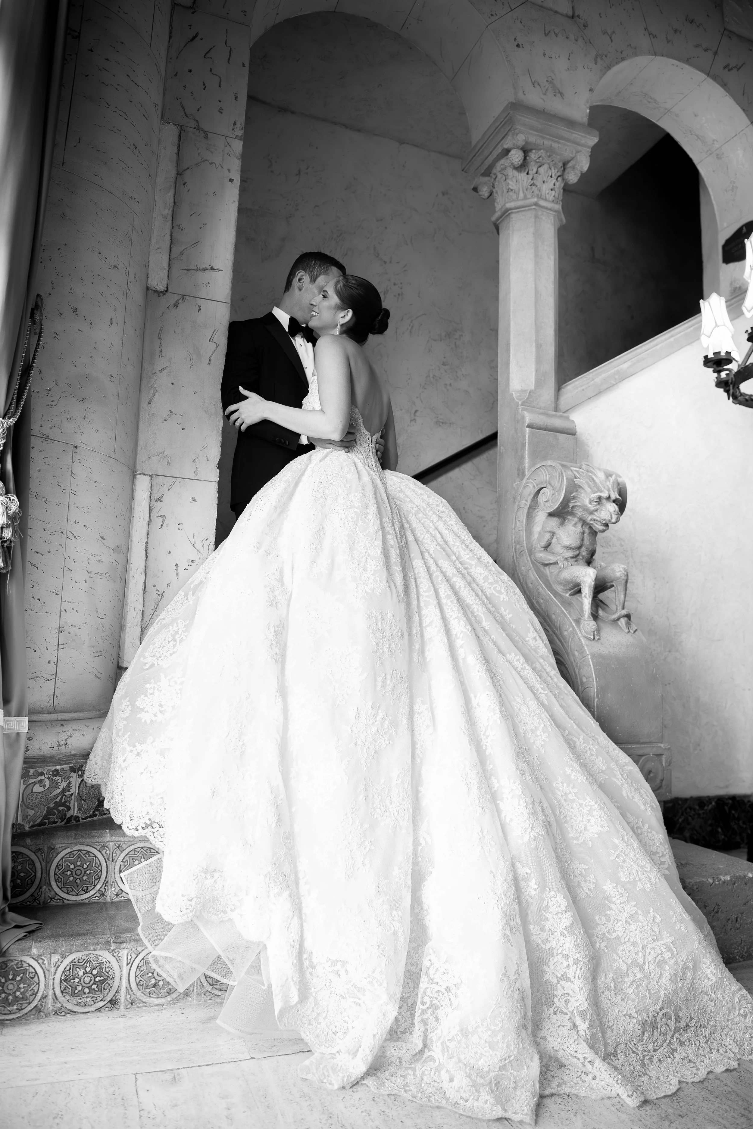 A bride and groom in wedding attire sharing a moment on a grand staircase inside a historic building with ornate architectural details.