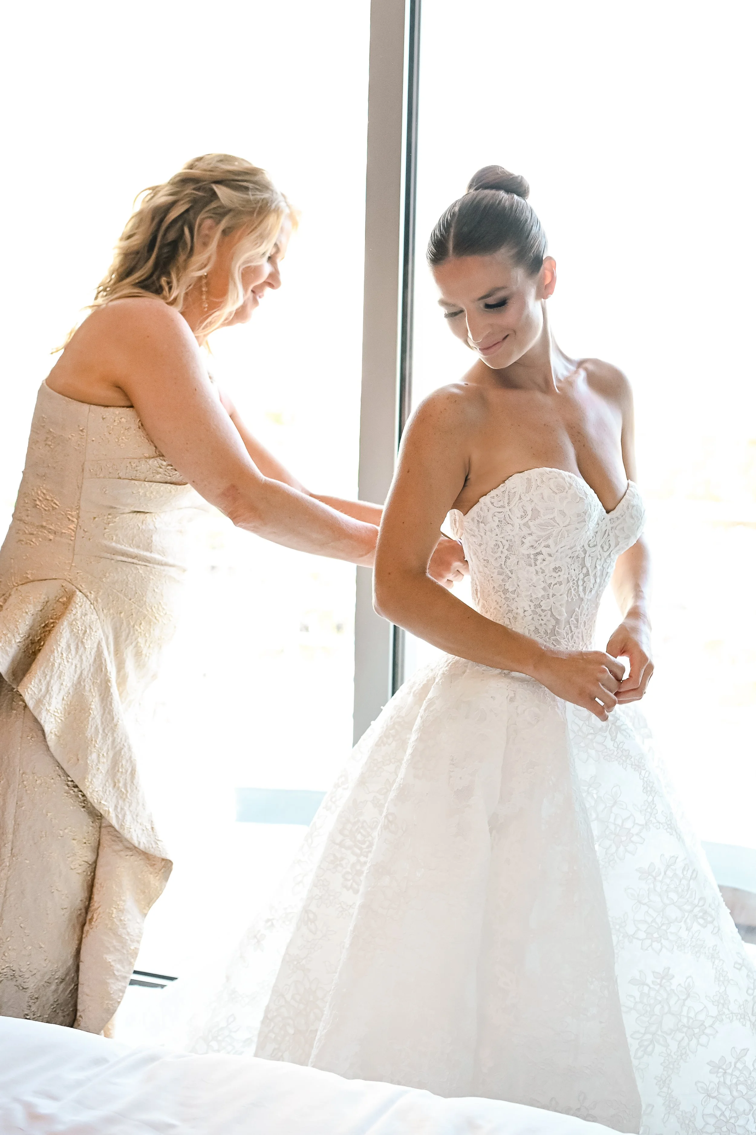 A bride in a strapless white wedding gown with lace details smiling as she gets ready, with another woman in a gold dress helping her.