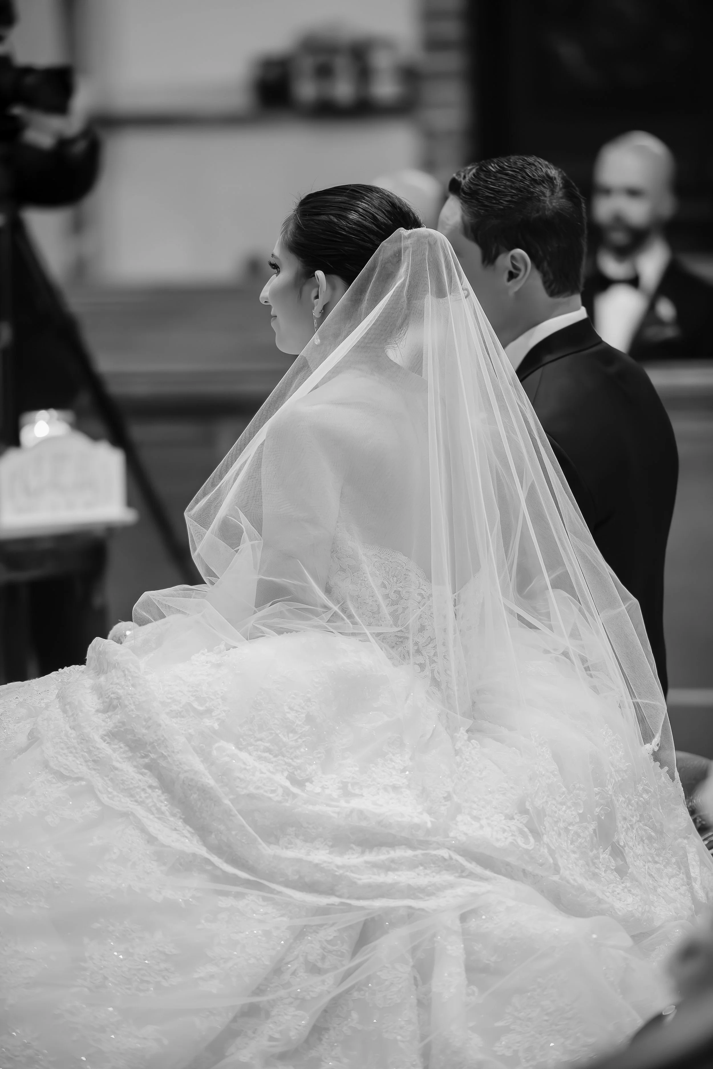 Black and white photo of a bride and groom standing side by side during a wedding ceremony, with the bride wearing a veil and lace gown, and the groom in a tuxedo.