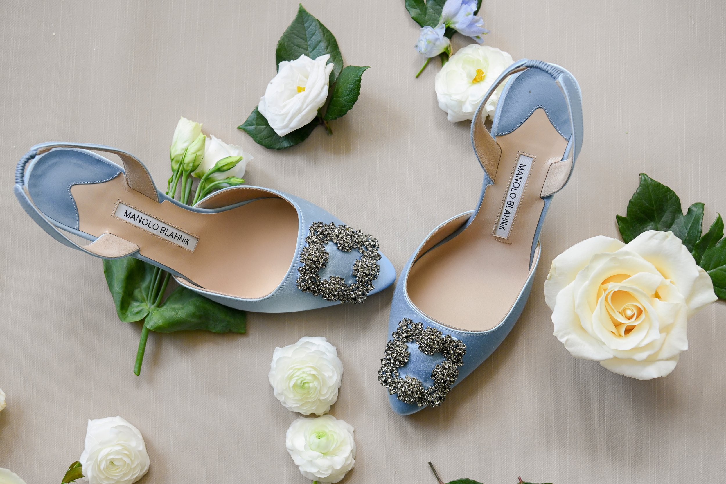 Light blue women's heels with rhinestone embellishments and a Manolo Blahnik label, surrounded by white flowers and green leaves on a beige background.
